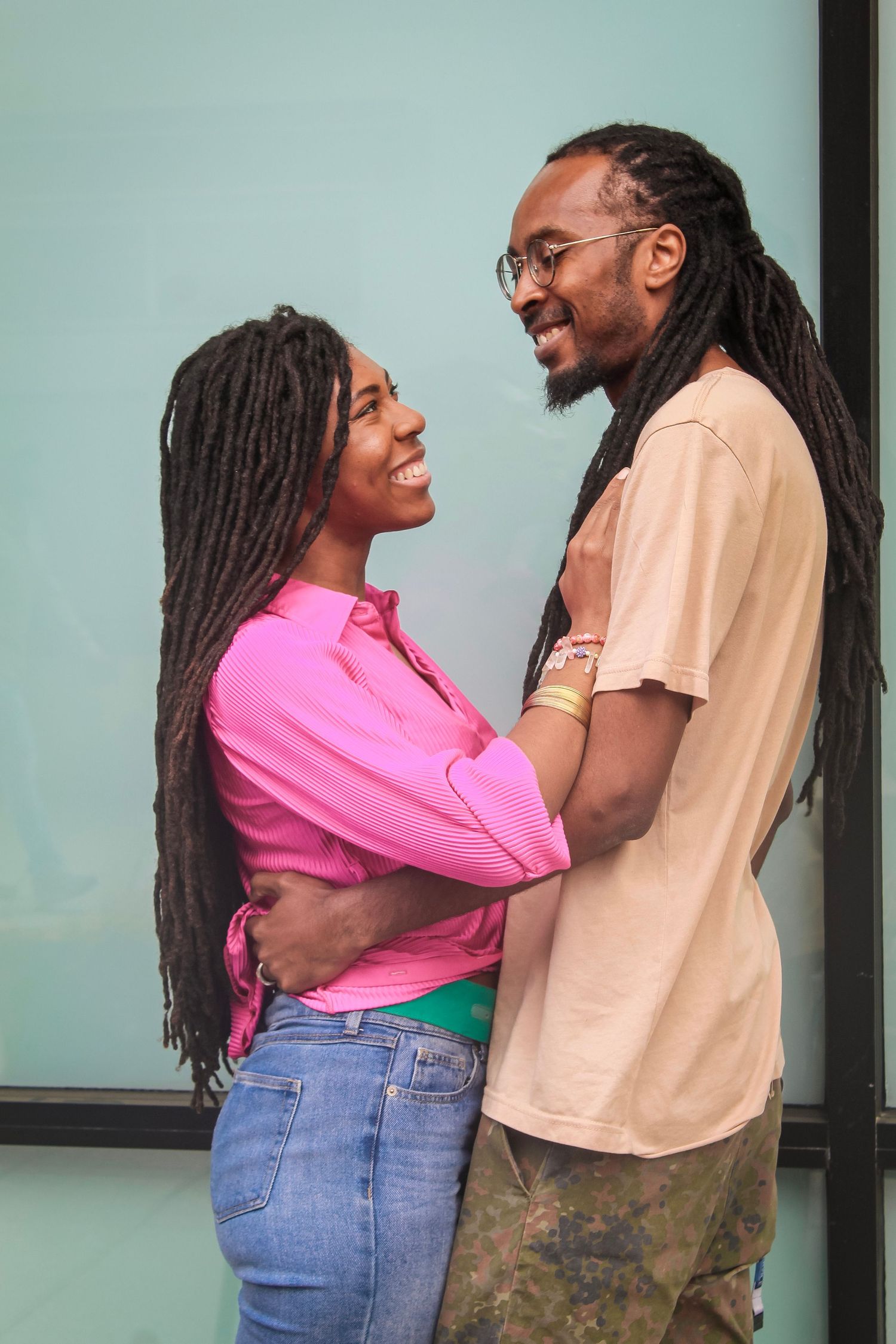 A loving couple with dreadlocks share a joyful moment together, wearing pink and beige shirts against a mint green wall.