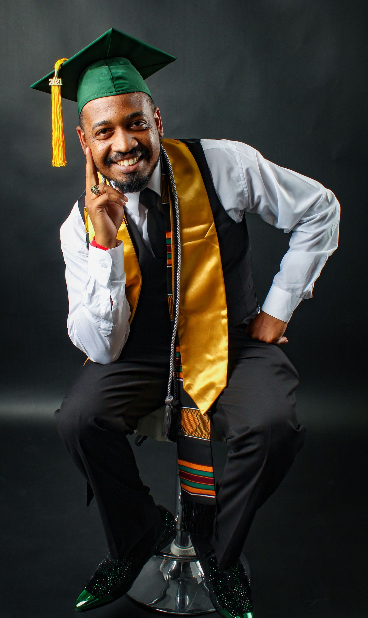 Graduate in black vest and gold stole poses against dark backdrop for professional portraits.