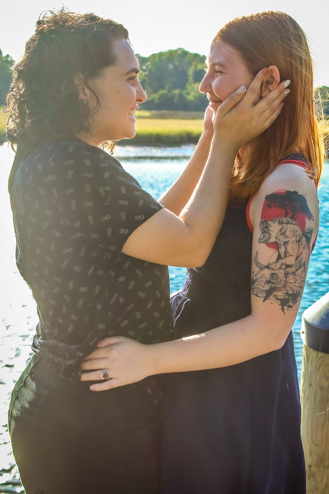 Two women share a tender moment by a waterfront, one cradling the other's face, with tattoos visible in warm sunlight.