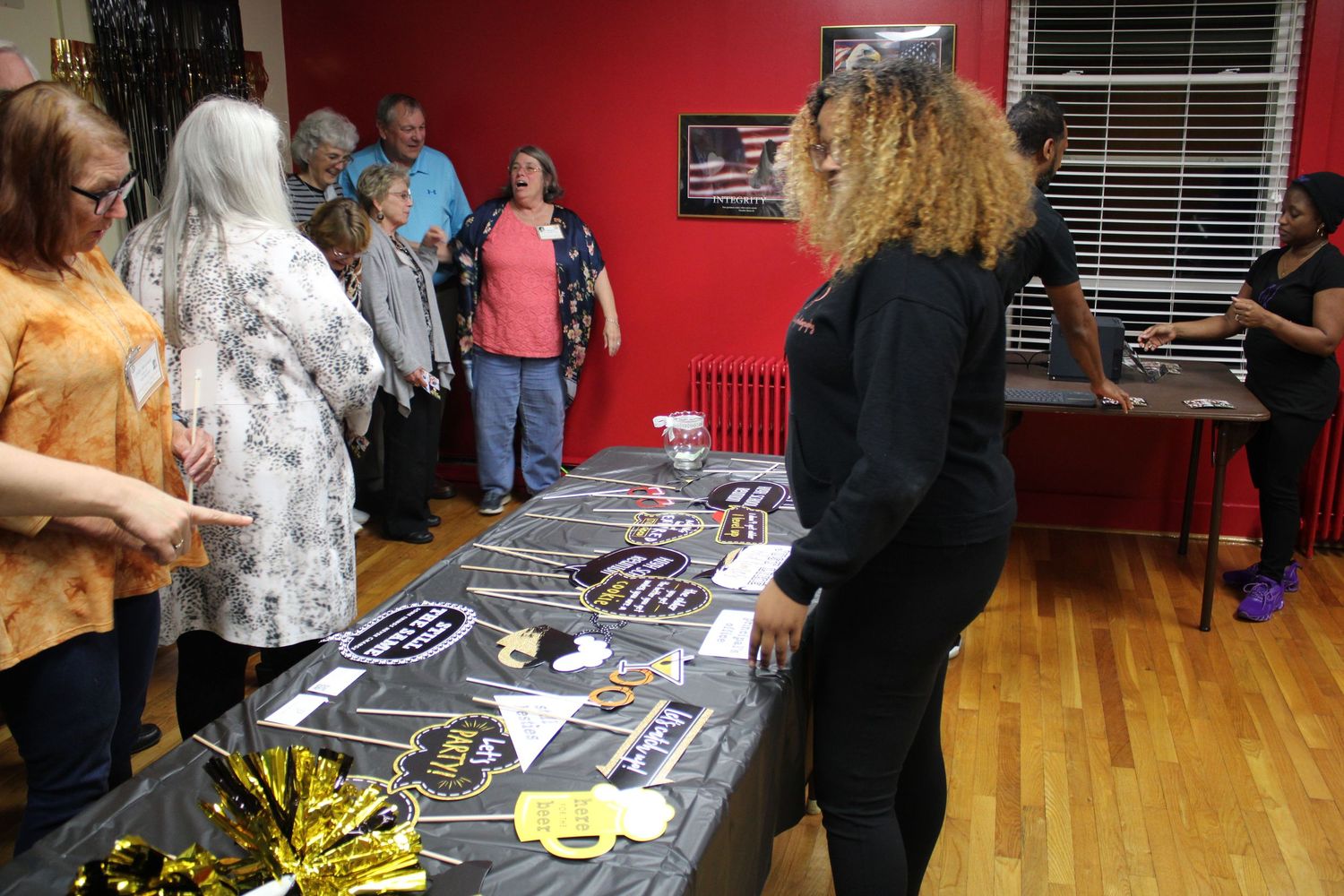 People gather around a merchandise table with items displayed at an indoor event with red walls.