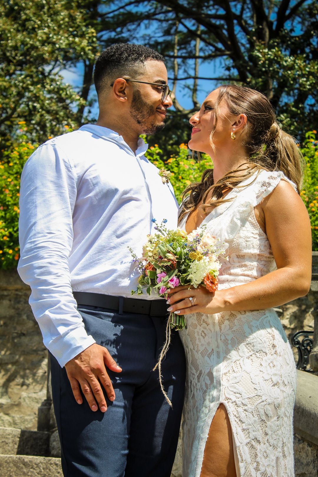 A bride and groom share a romantic moment outdoors, she holds a wildflower bouquet in a lace dress as he wears a white shirt.