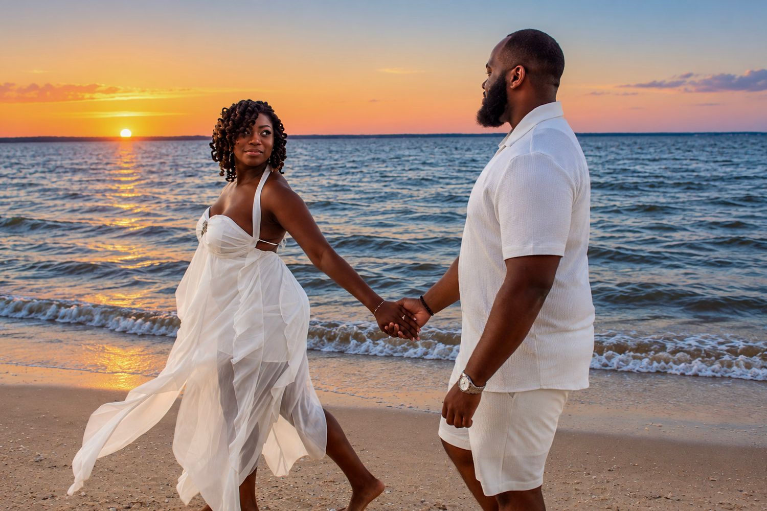 Couple in white outfits holding hands on a beach at sunset, waves gently lapping the shore behind them.
