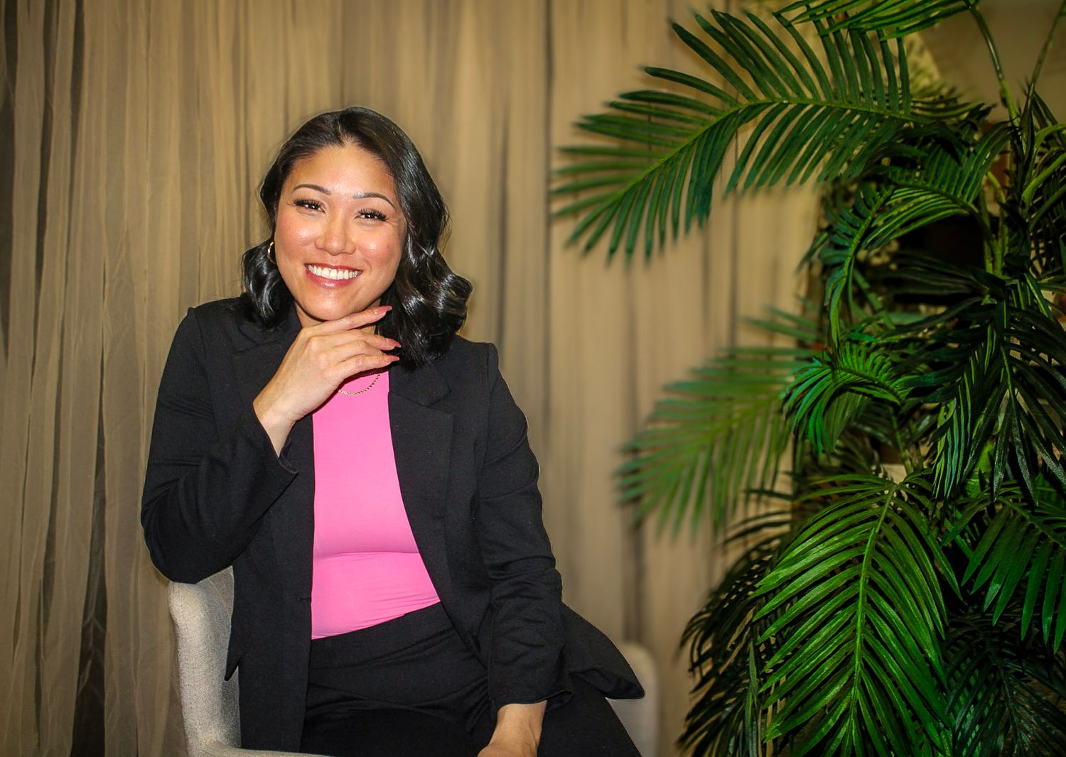 Smiling woman in pink top and black blazer poses near tropical palm plant against beige curtain backdrop.