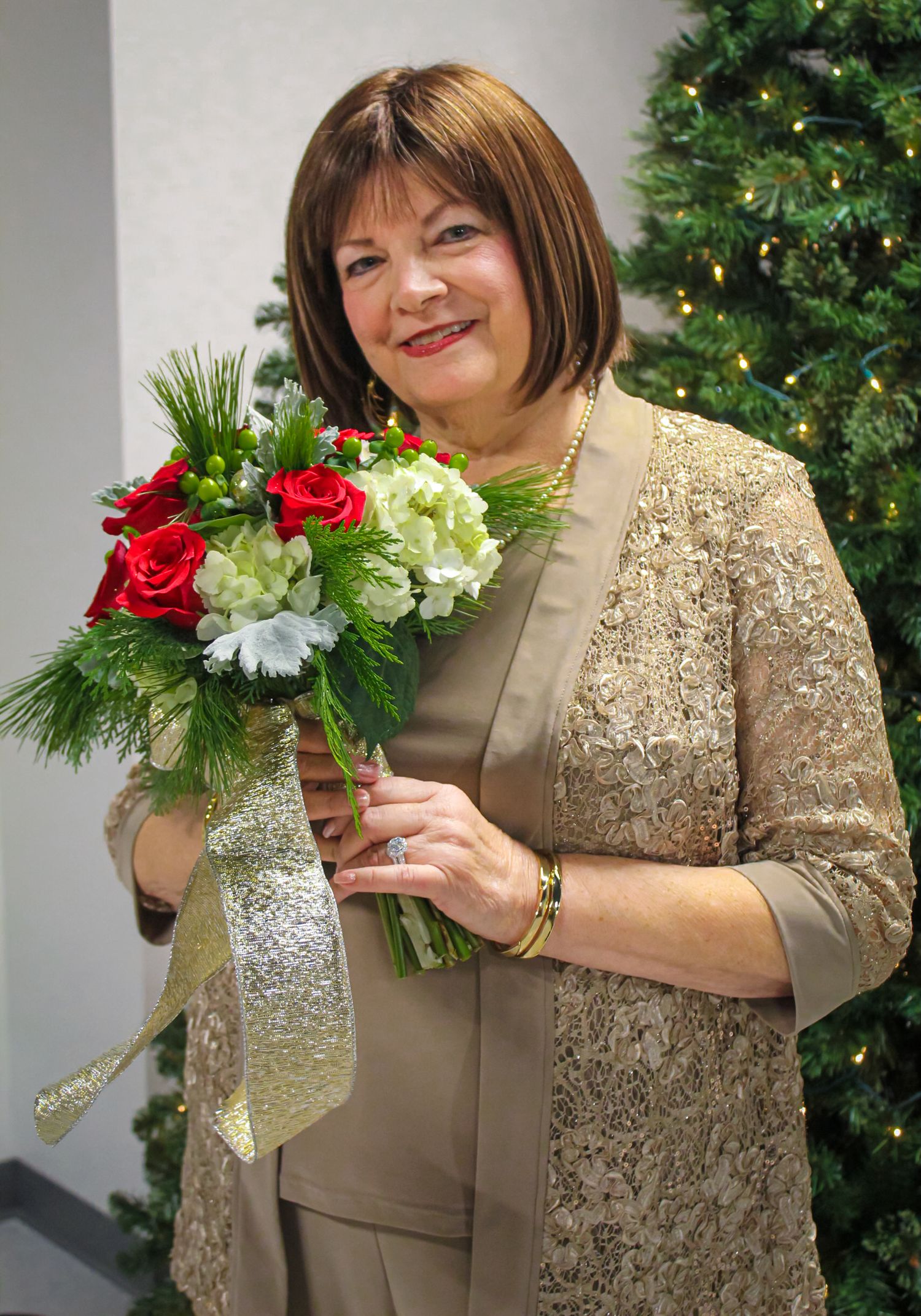 Woman in gold lace jacket holding red roses and white hydrangea bouquet near a decorated Christmas tree.