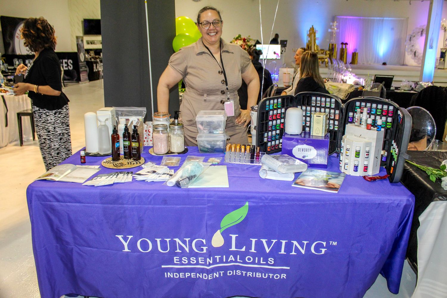 Woman smiling behind a Young Living Essential Oils Independent Distributor booth at an indoor vendor event.