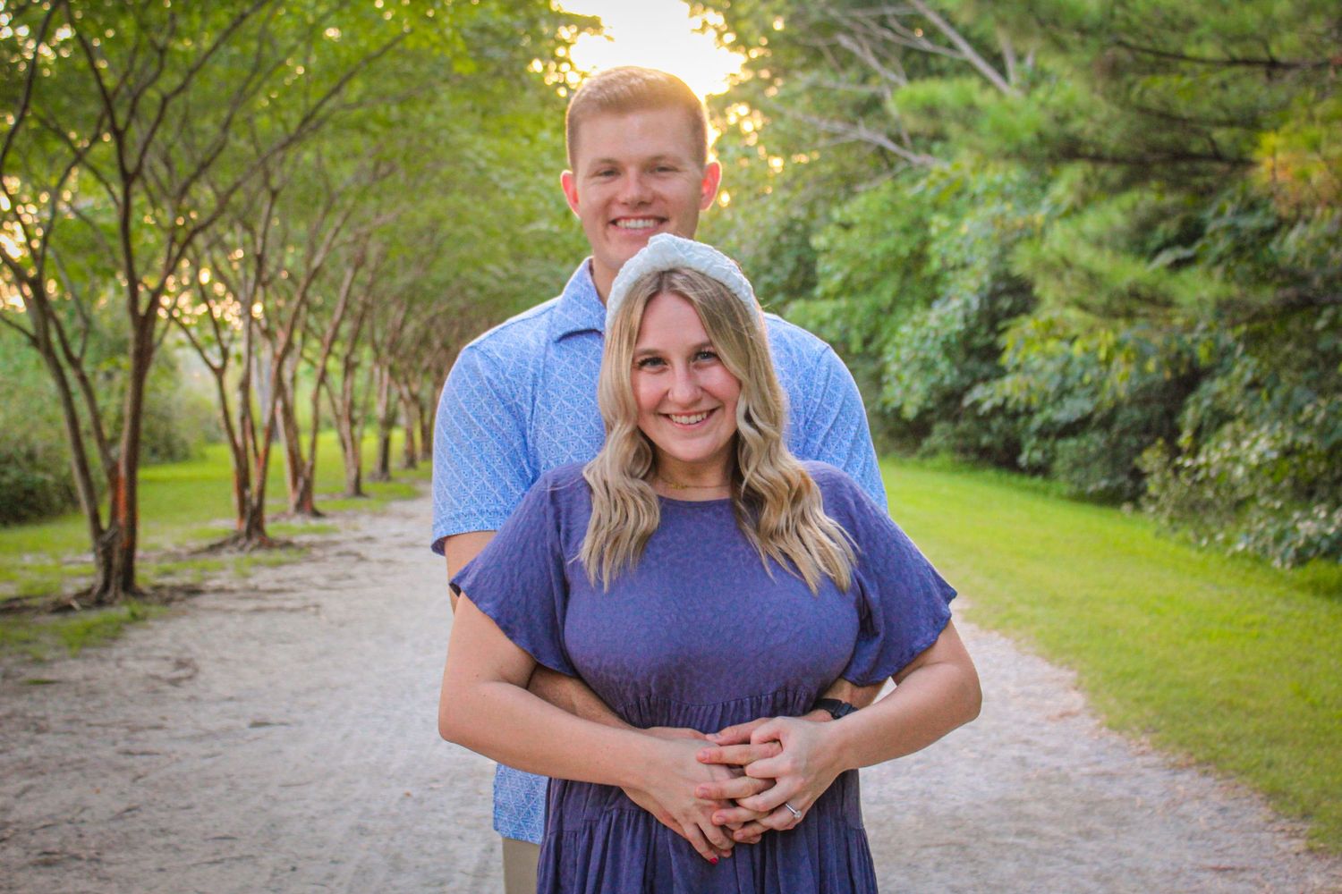 A smiling couple poses on a tree-lined path, woman in purple dress, man in blue shirt, golden hour lighting.