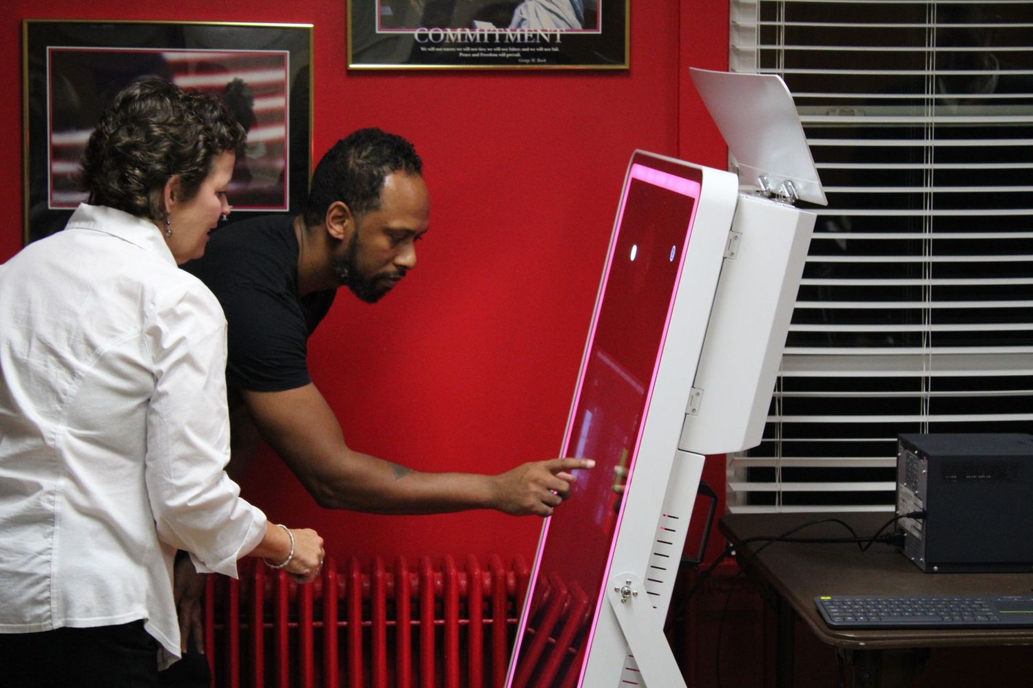 Two people examine equipment in front of a red wall with window blinds.