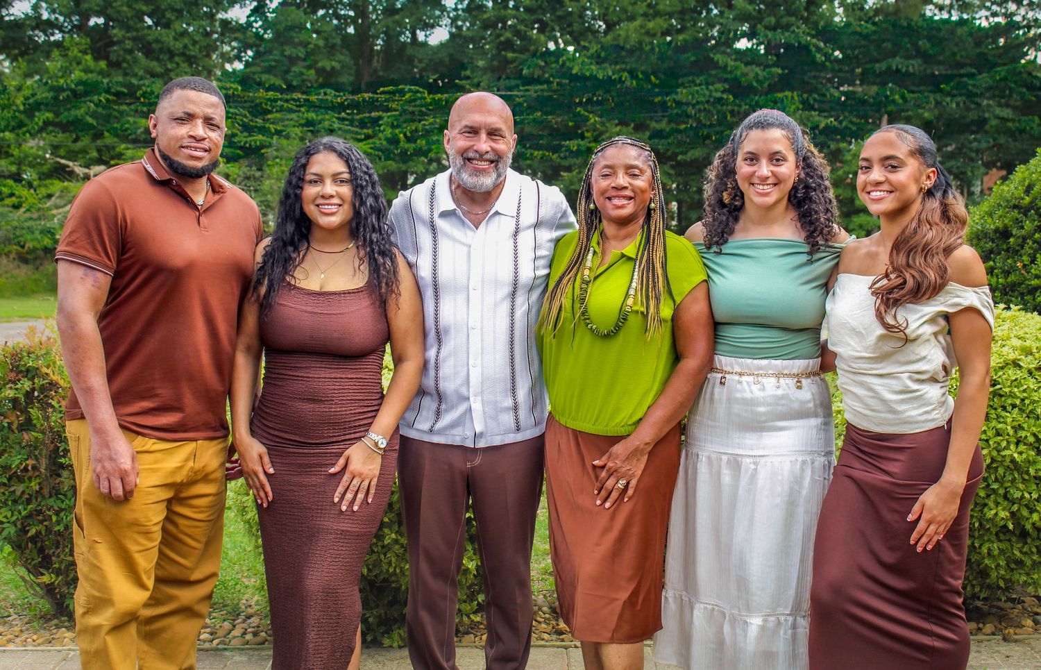 A joyful family of six poses together outdoors, smiling in coordinated brown and green outfits.