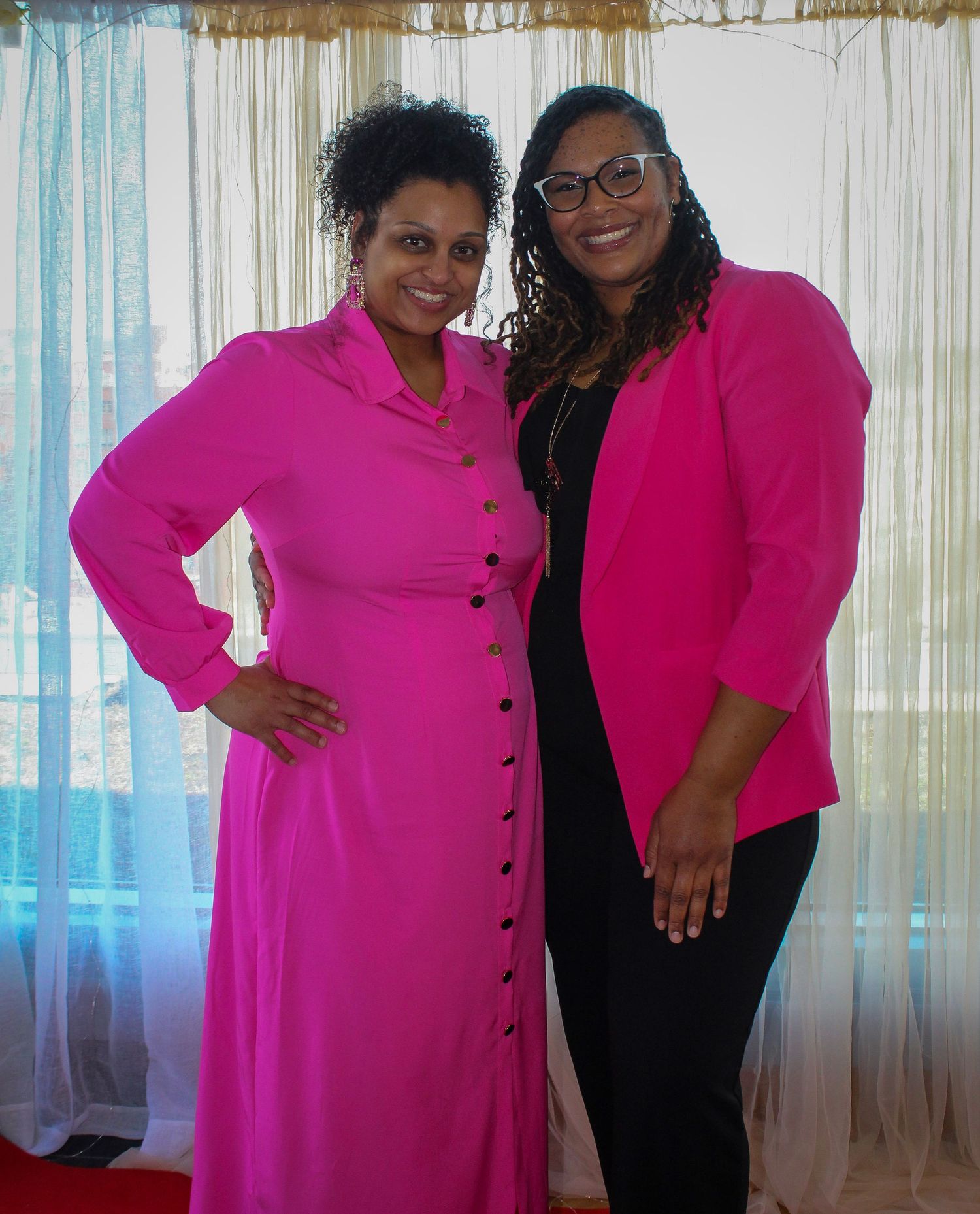 Two women in matching pink outfits smiling together at a formal event indoors.