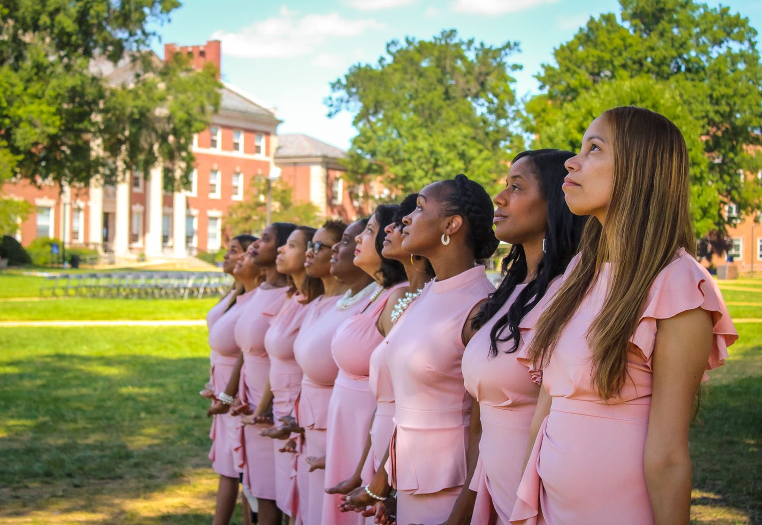 A line of people in coordinated pink outfits pose in front of a stately brick building.