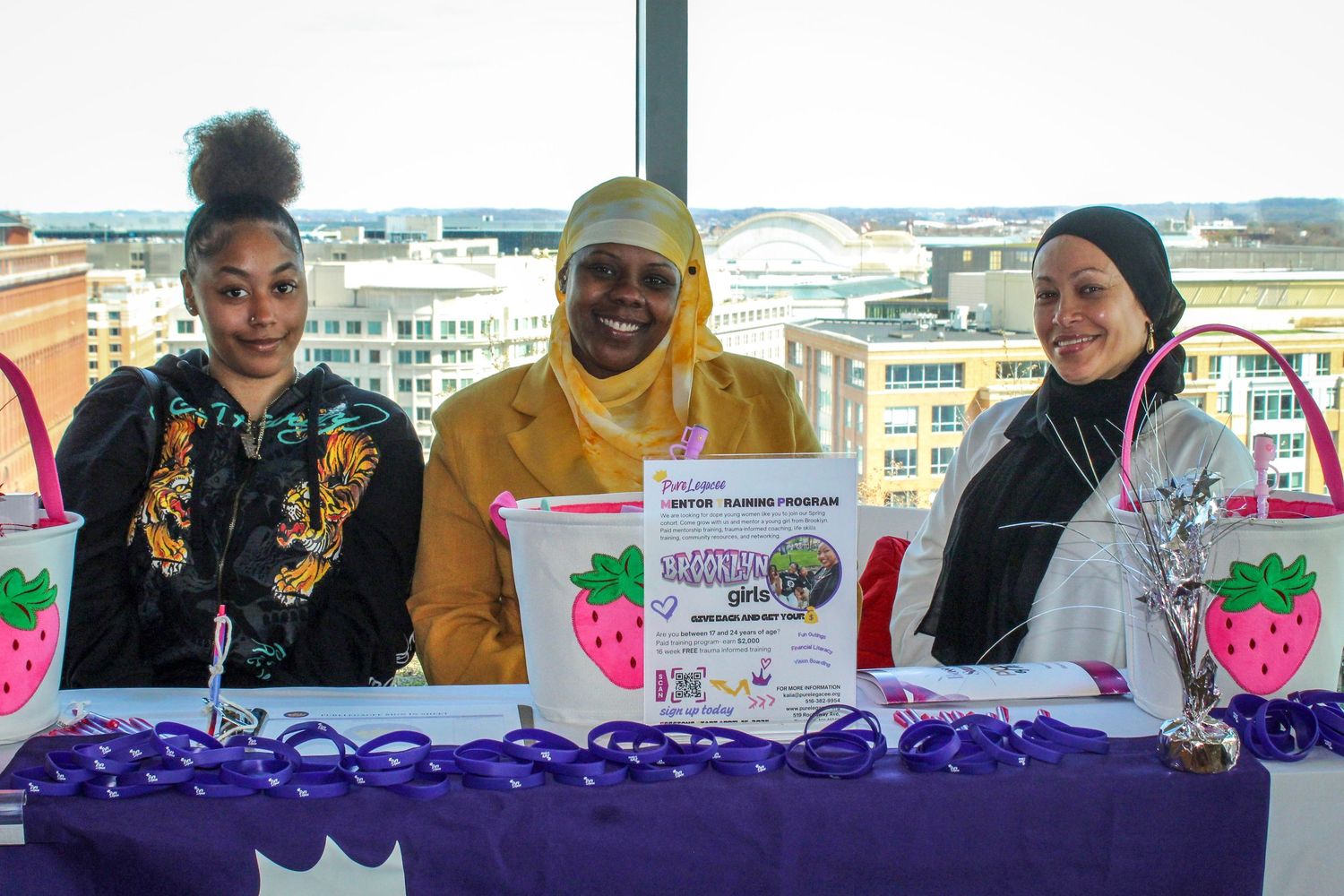 Three women smile at a purple-draped event table with strawberry-themed decorations and promotional signage indoors.