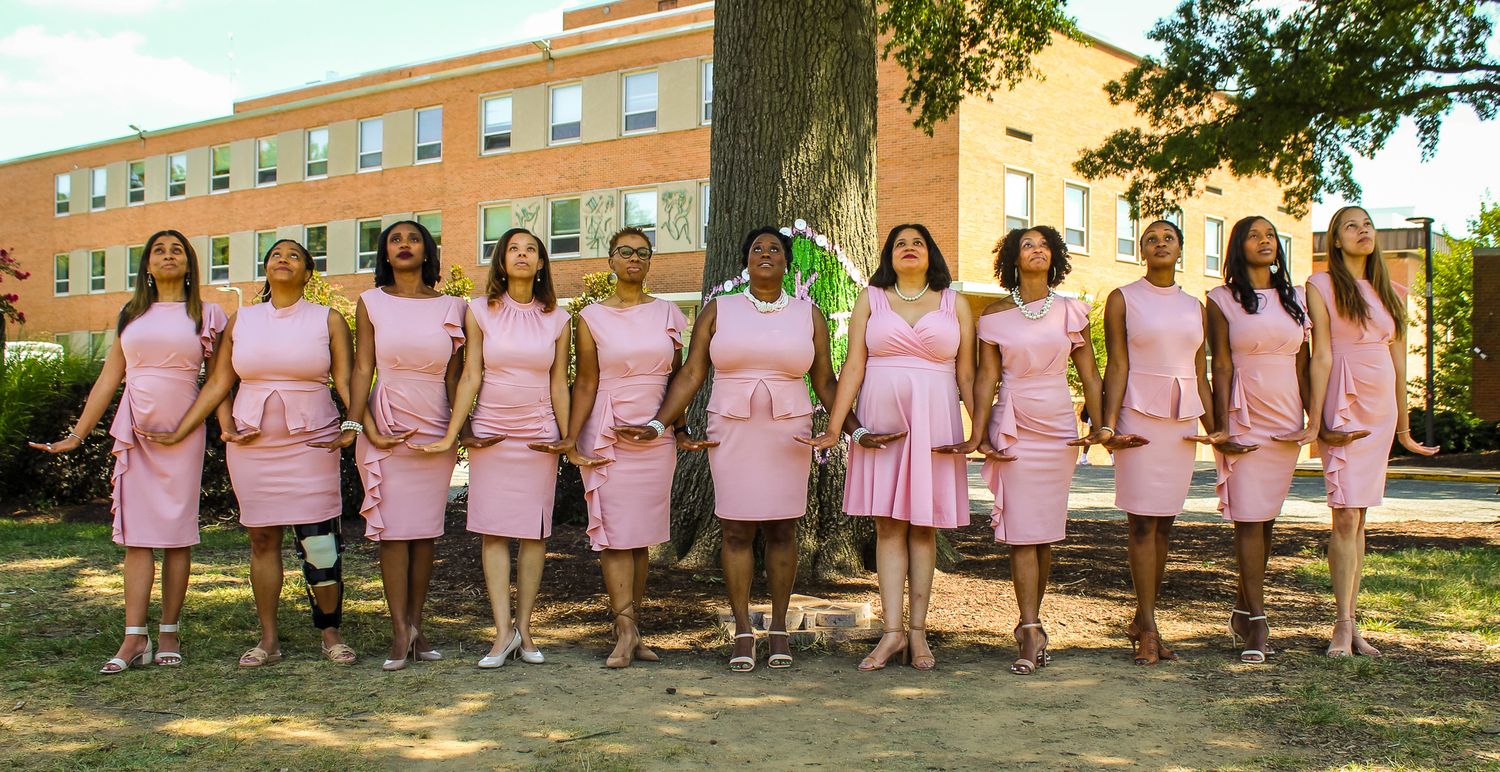 Group of women in matching pink dresses posing outdoors on a sunny day near a brick building.