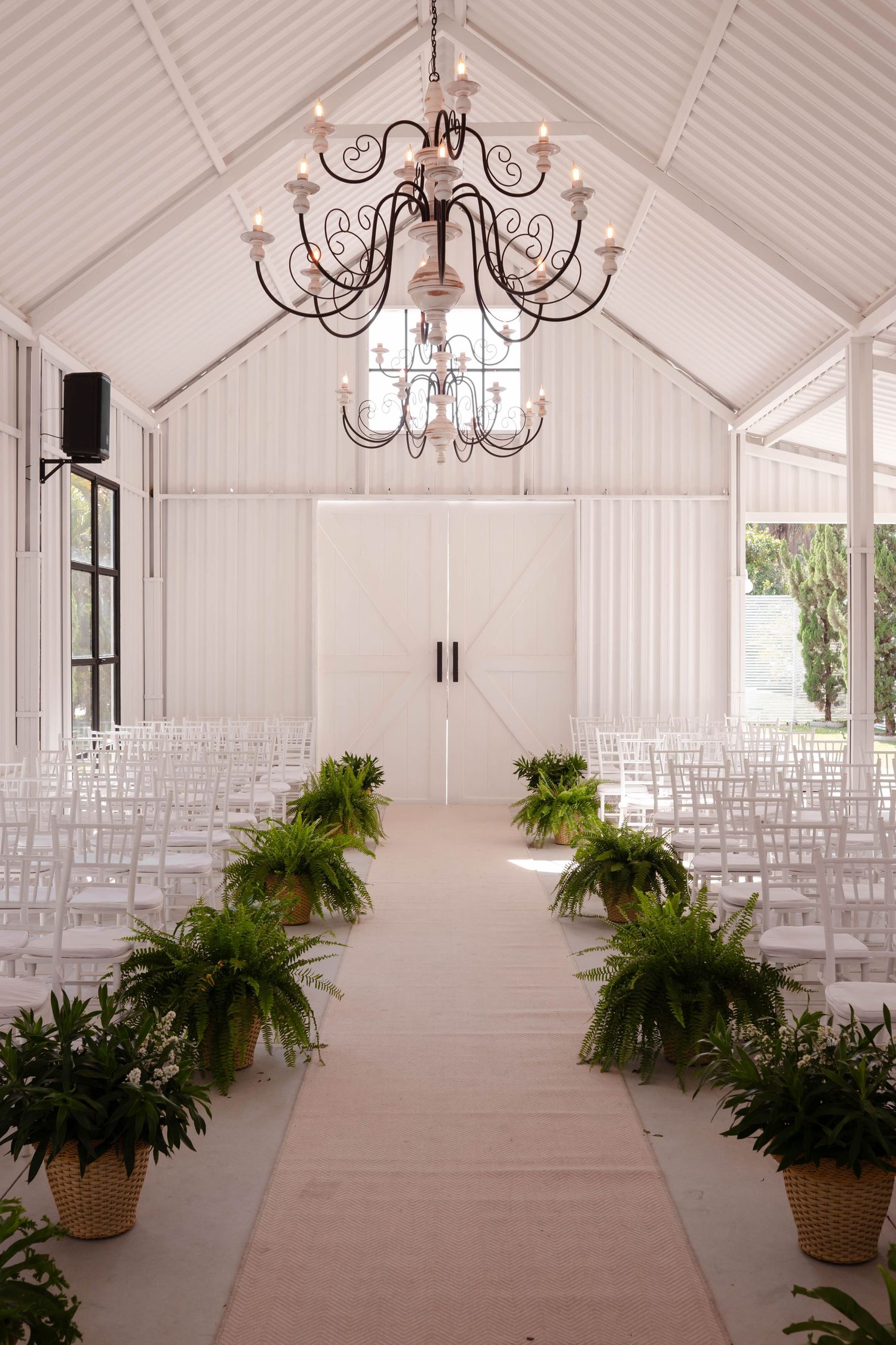 White wedding chapel interior with rows of chairs, chandeliers, and potted ferns creating an elegant atmosphere.