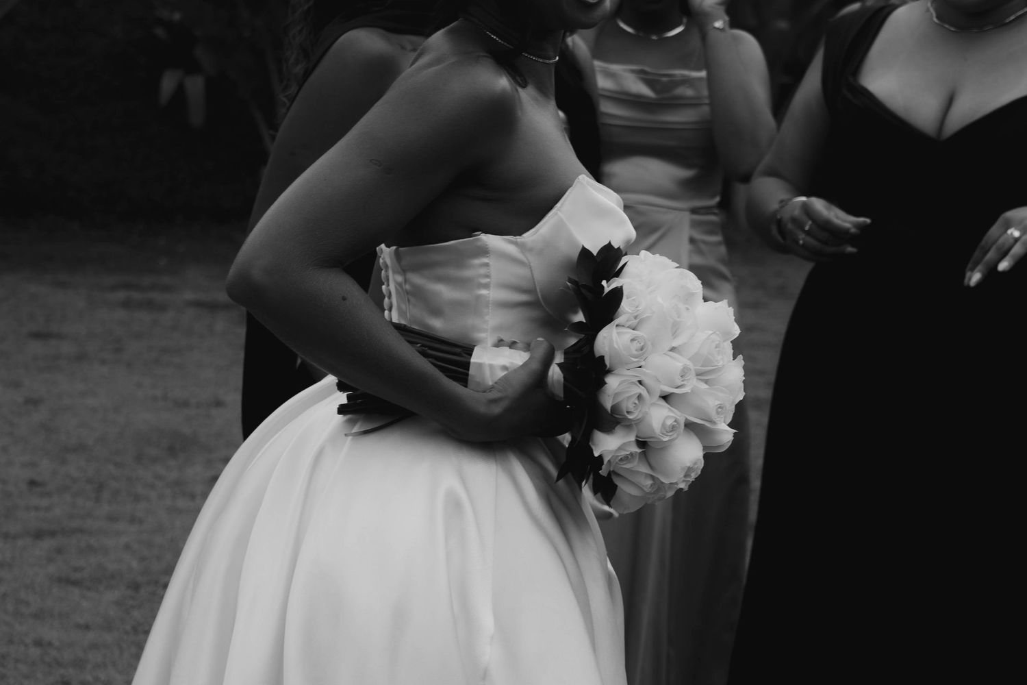 A wedding party poses in a line outdoors with bridesmaids in dark dresses and a bride in a white gown holding bouquets.