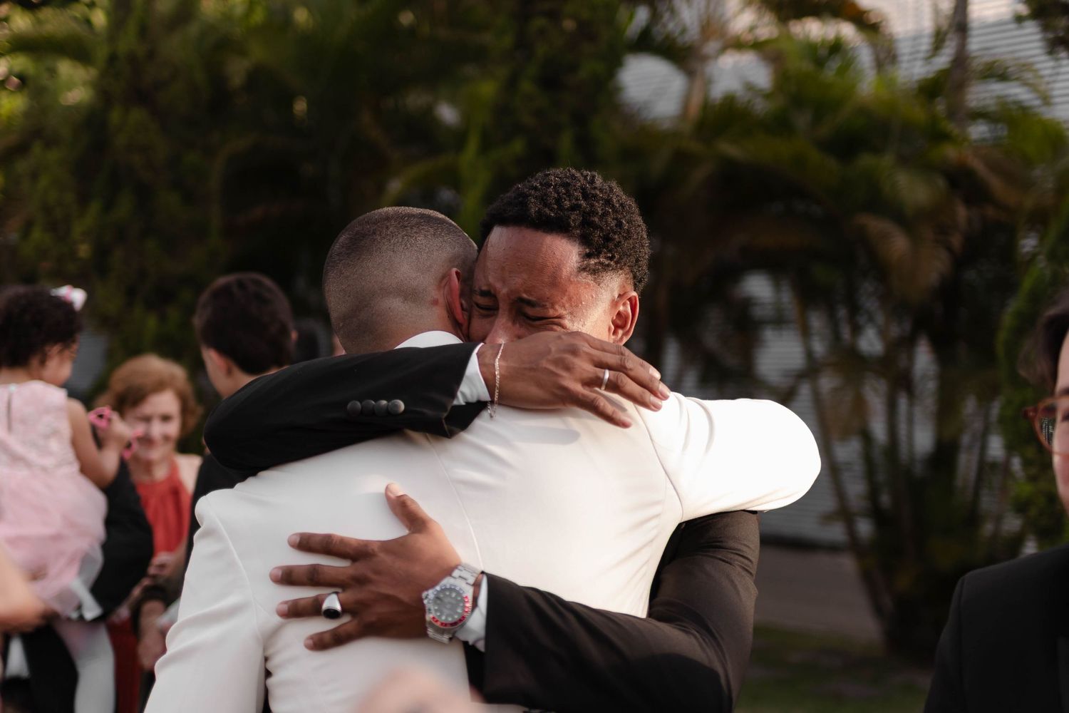 Two people share an emotional embrace at an outdoor gathering with palm trees in the background.