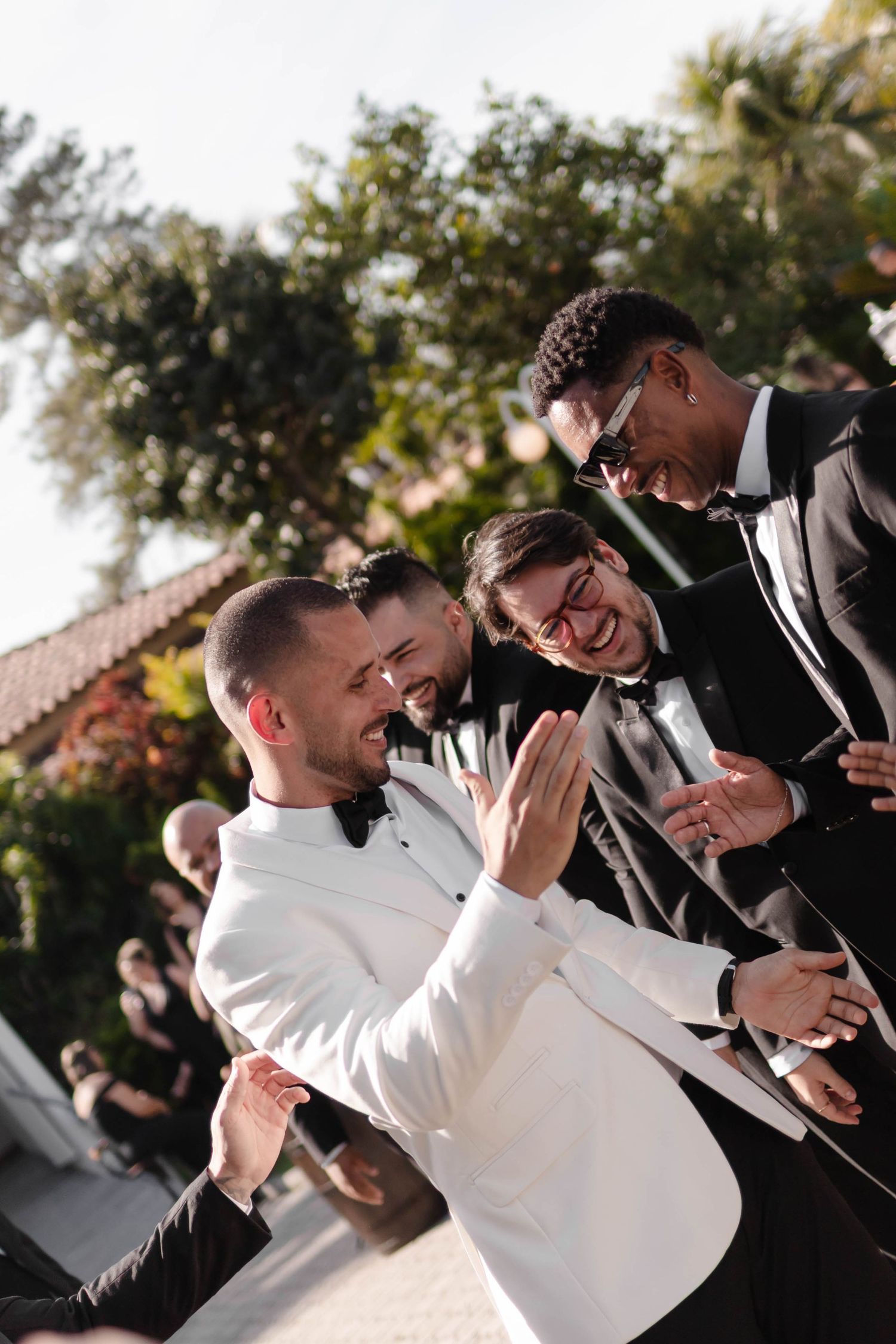 A group of groomsmen in black suits share a candid laughing moment with someone in a white jacket at an outdoor event.