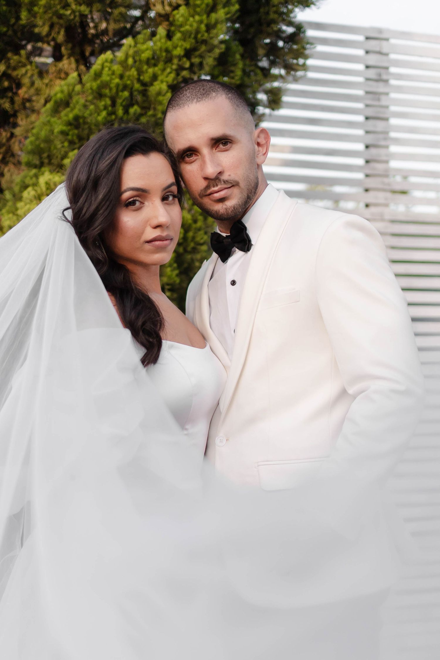 Romantic wedding portrait series showing couple in white attire against white shutters with flowing veil and natural lighting.