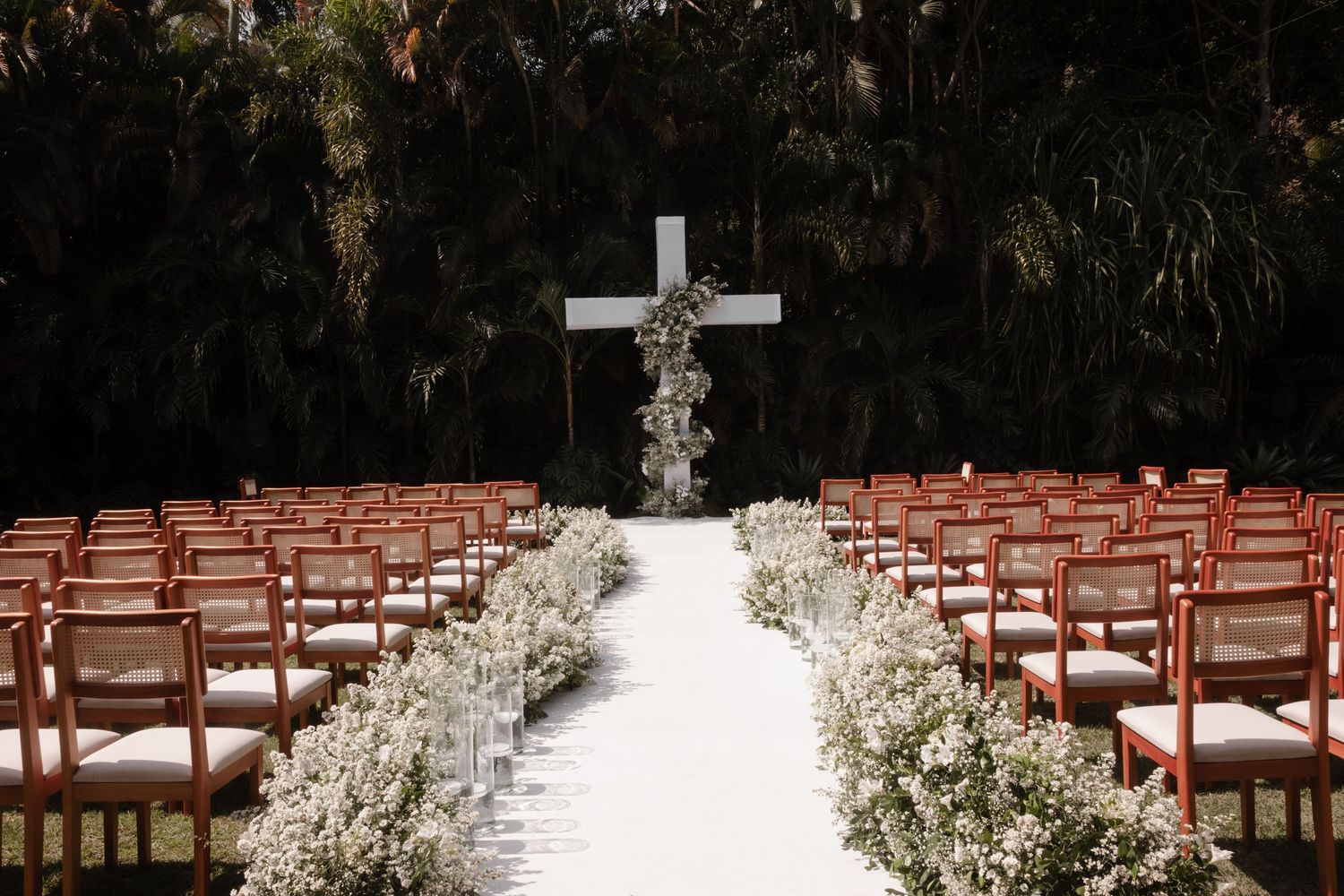 White floral arrangements line an outdoor wedding aisle with wooden chairs and a cross altar under tree canopy.