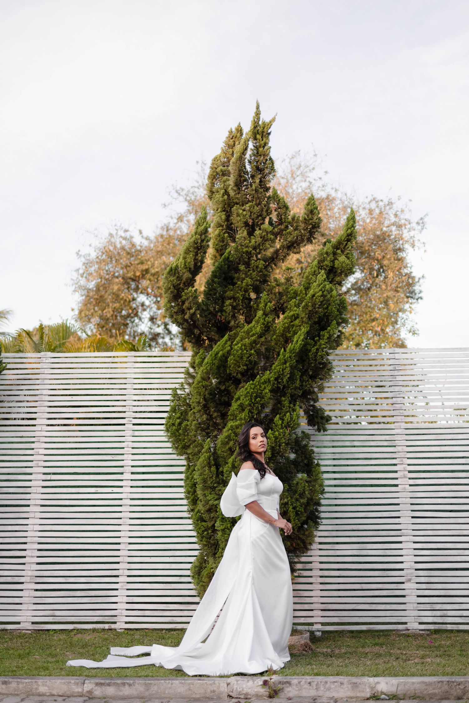 Wedding photography series featuring a bride posing against a white slatted fence and cypress trees.
