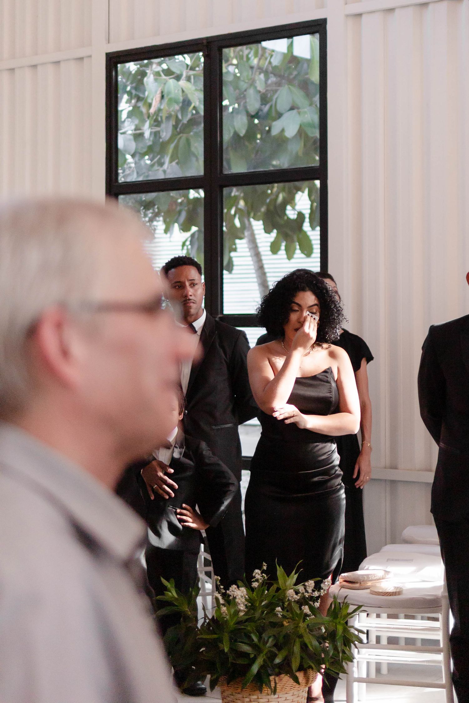 People in formal black attire gather at an indoor event space with white curtains and plants in the background.