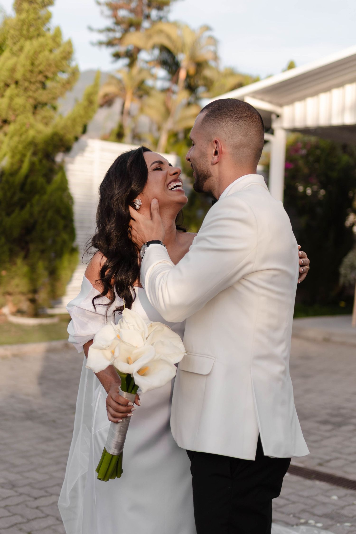Newlyweds in white attire sharing romantic kiss during outdoor wedding photoshoot.