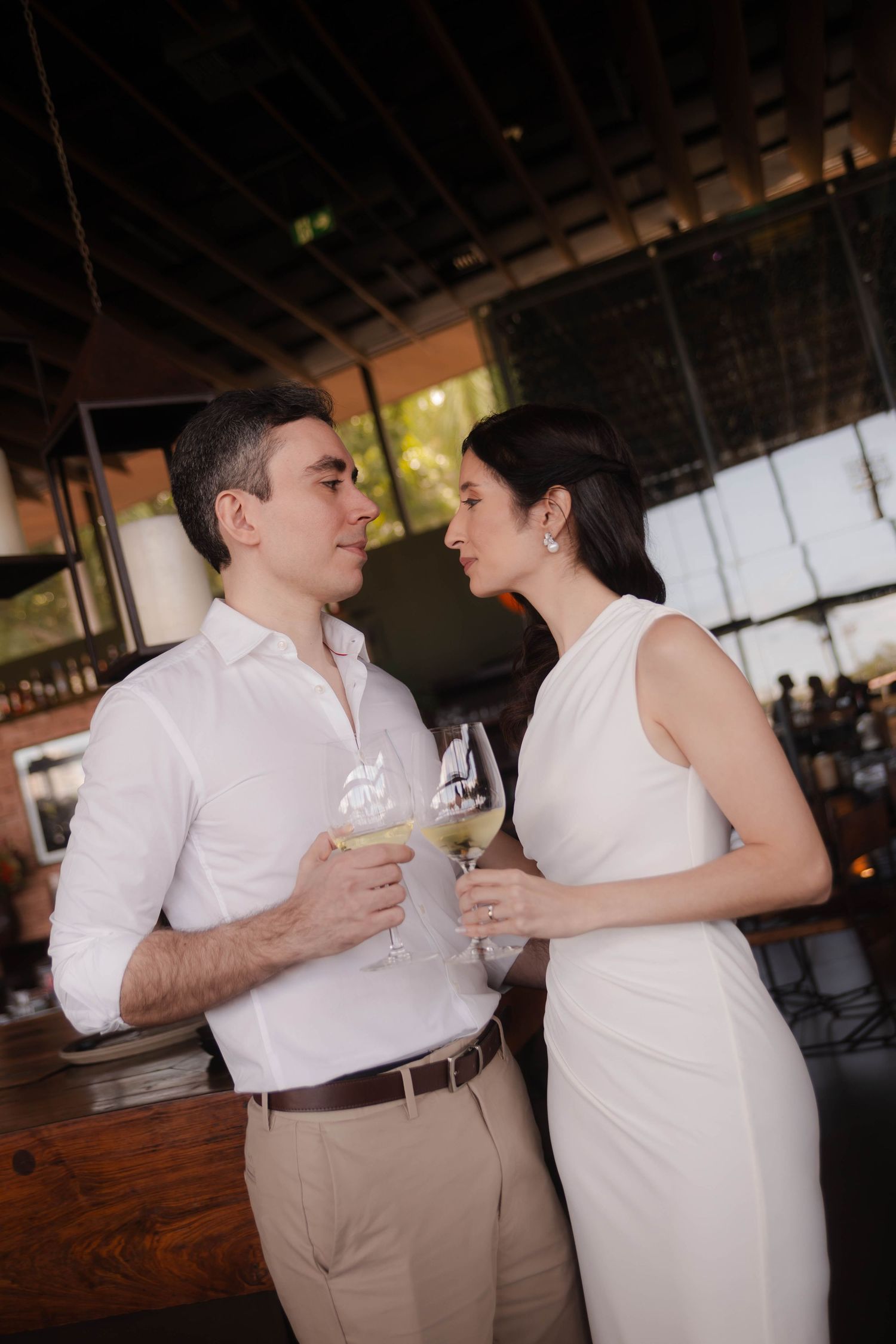 Couple in white attire sharing drinks at rustic wooden bar.