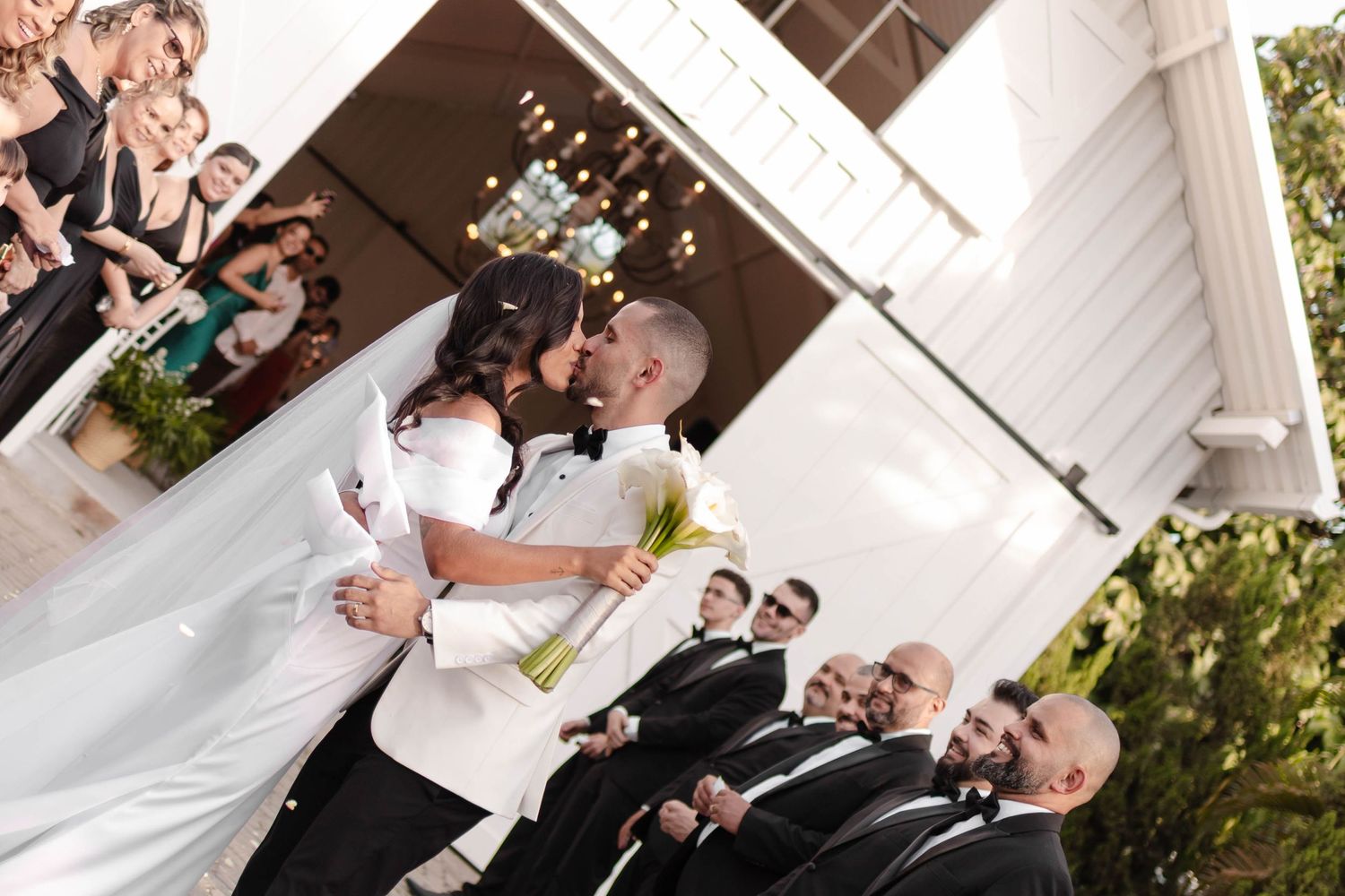 A couple shares their first kiss as newlyweds at an outdoor wedding ceremony while guests look on.