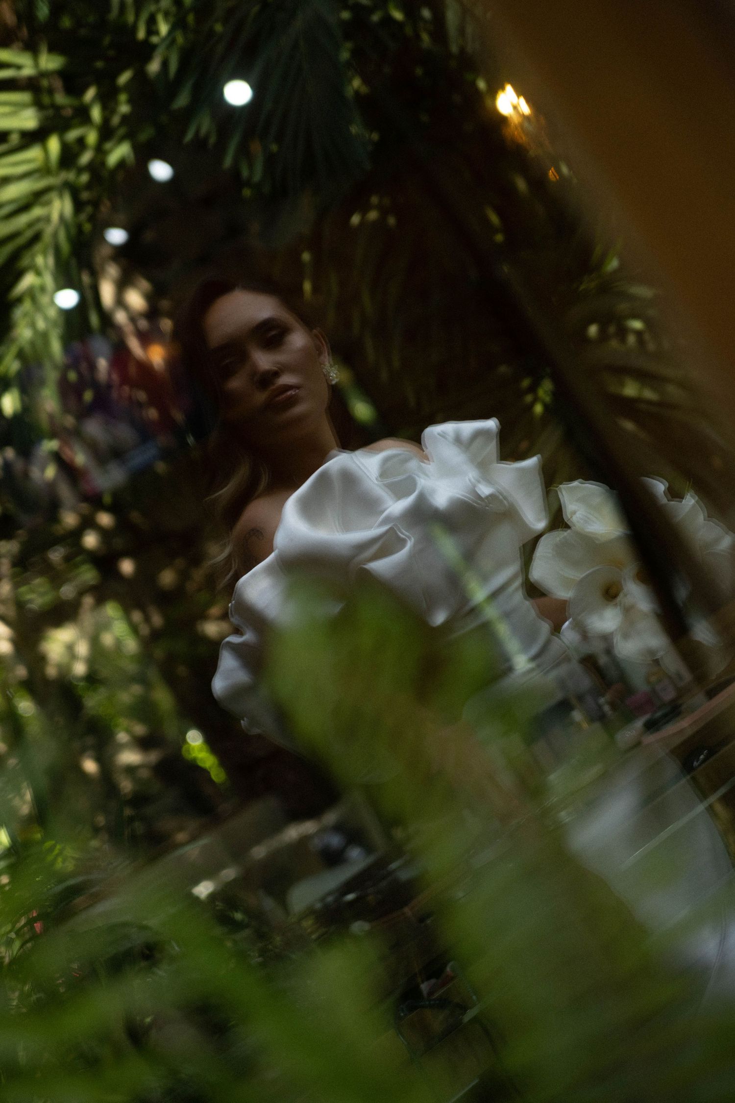 Ethereal wedding scene through greenery with white dress visible in soft focus.