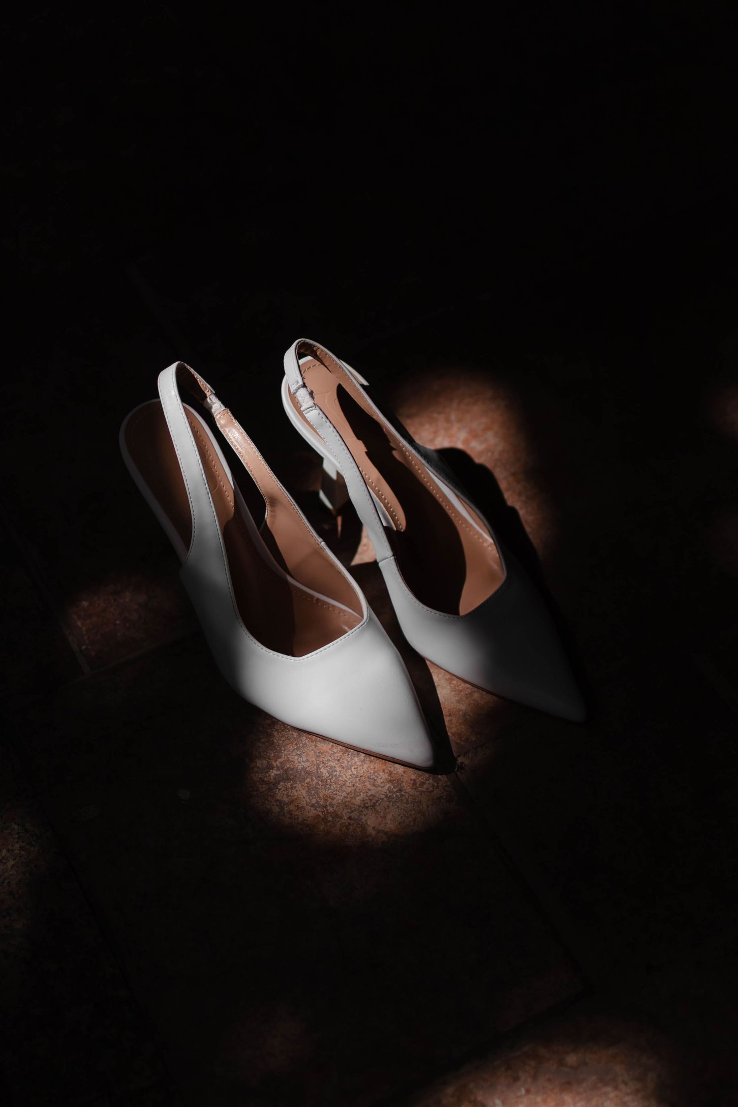 Elegant white slingback heels with pointed toes photographed dramatically against a dark background with spotlight effect.