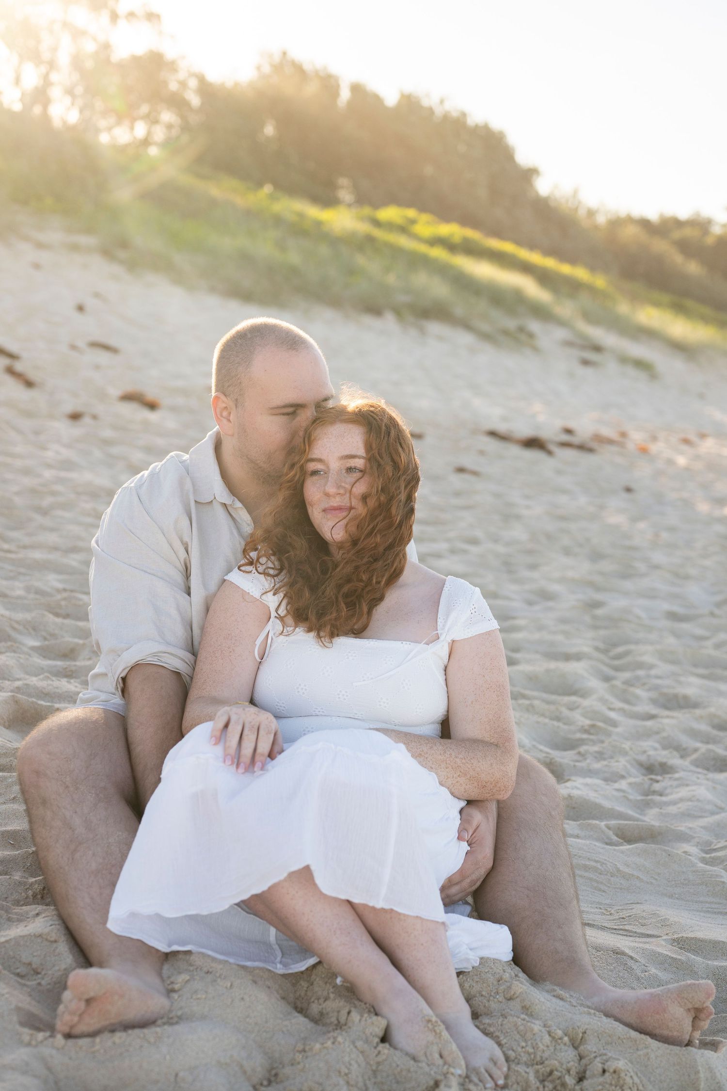 Couple sitting together on sandy beach at sunset, woman in white dress leaning back against man in casual linen shirt.