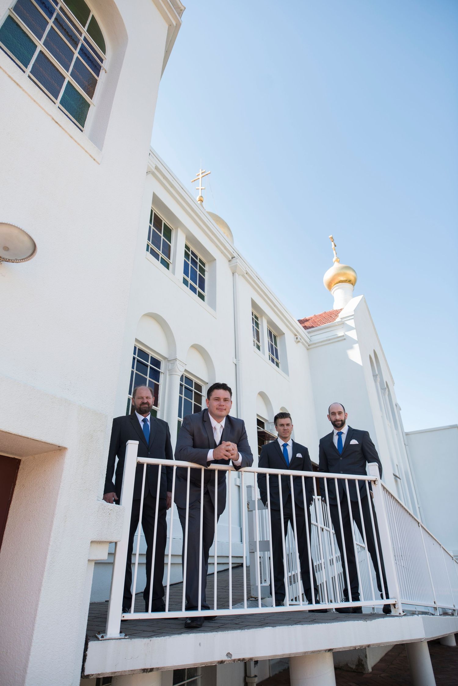 Several people in formal attire stand along a white balcony railing outside a Russian Orthodox Church.