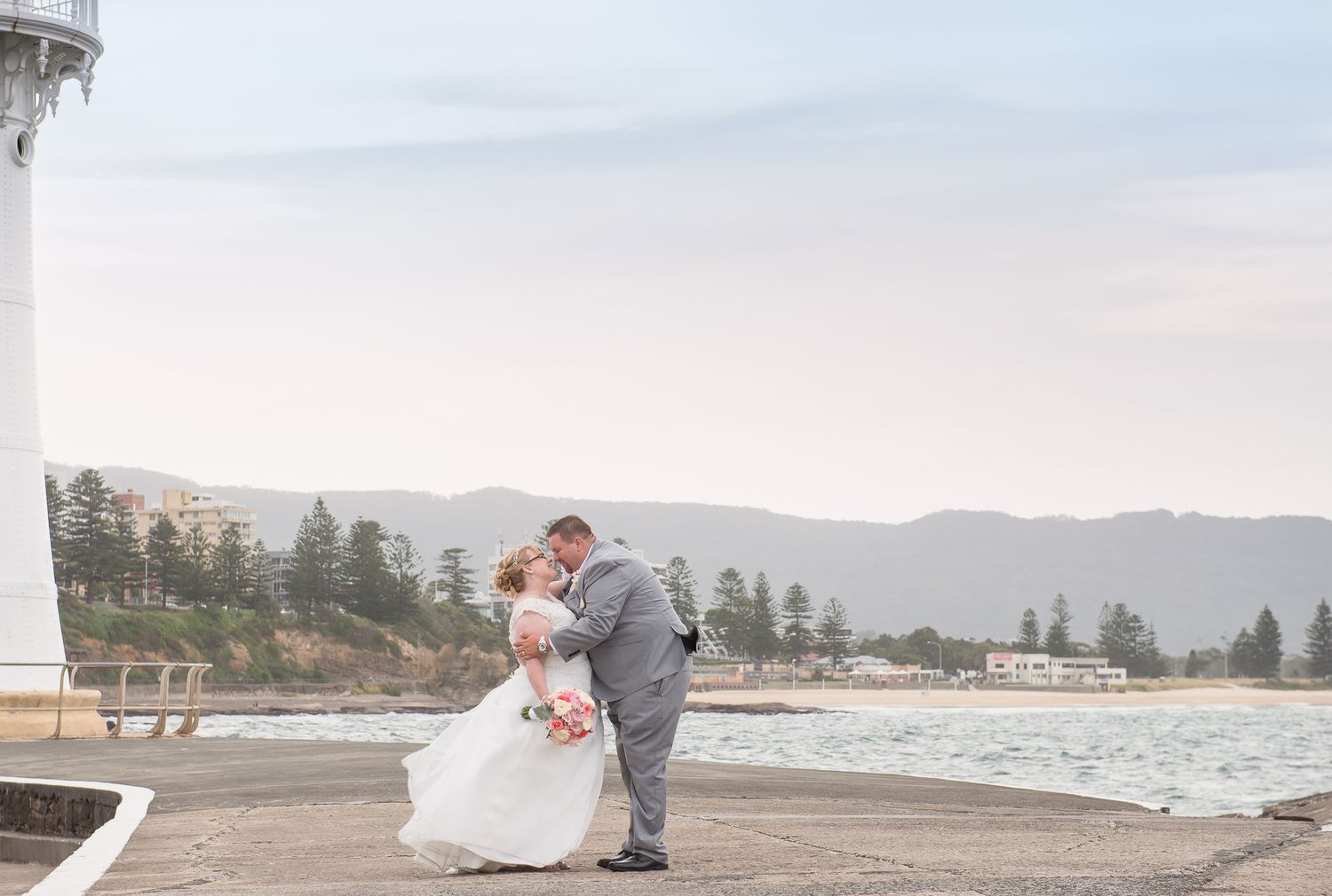 A couple in wedding attire embraces on a beach with mountains and lighthouse in the background.