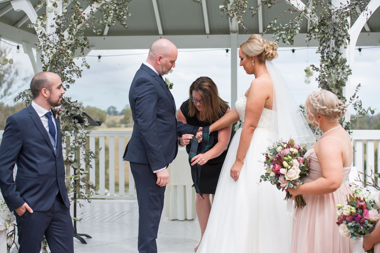Emotional Scottish hand fasting moment during an outdoor wedding ceremony under a white gazebo with floral decorations.