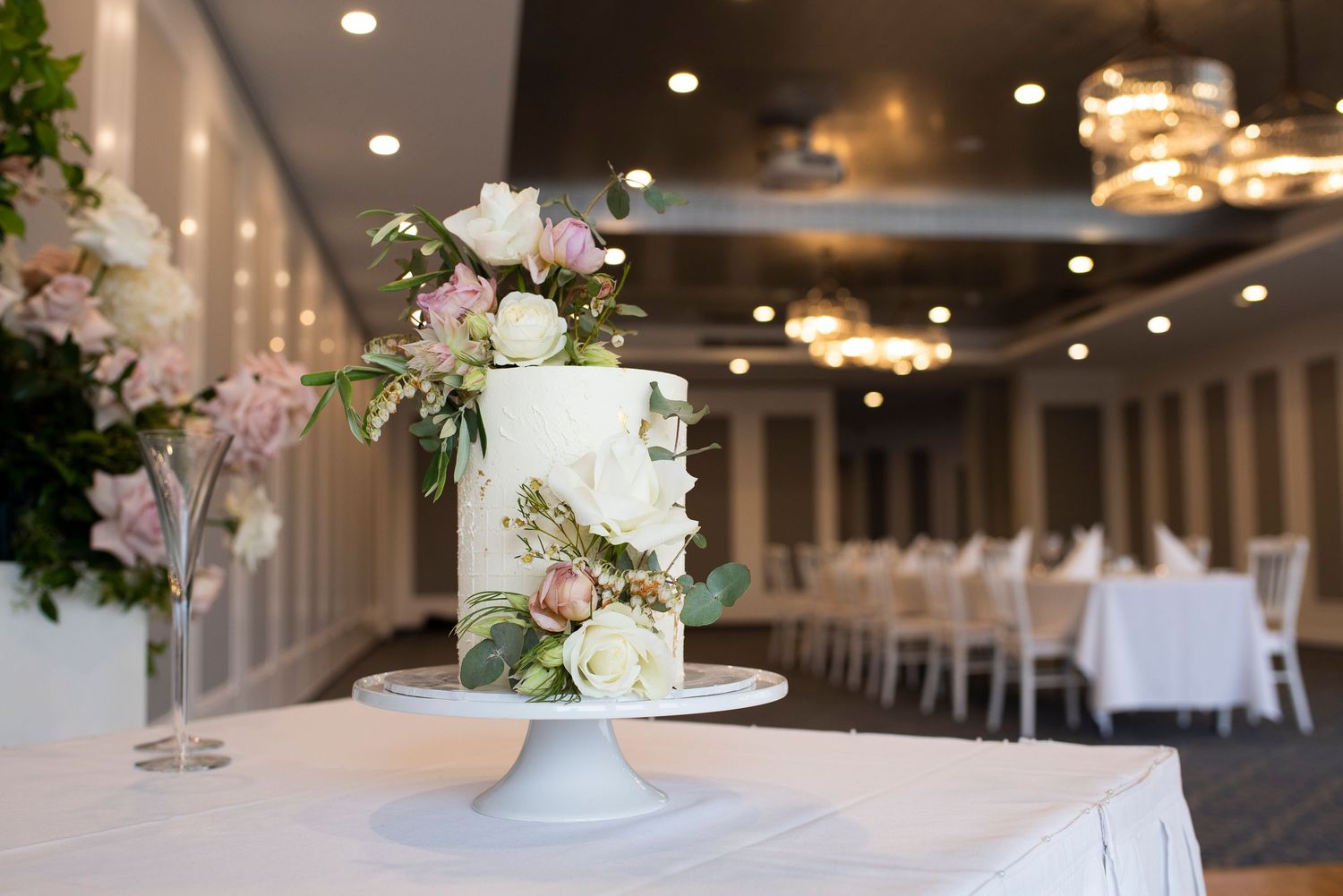 Elegant white wedding cake with floral decorations displayed on a pedestal in a reception venue.