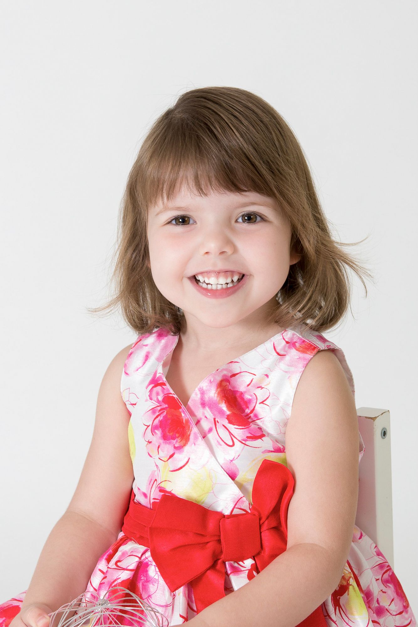 Child in floral dress with red bow smiles brightly against white background.