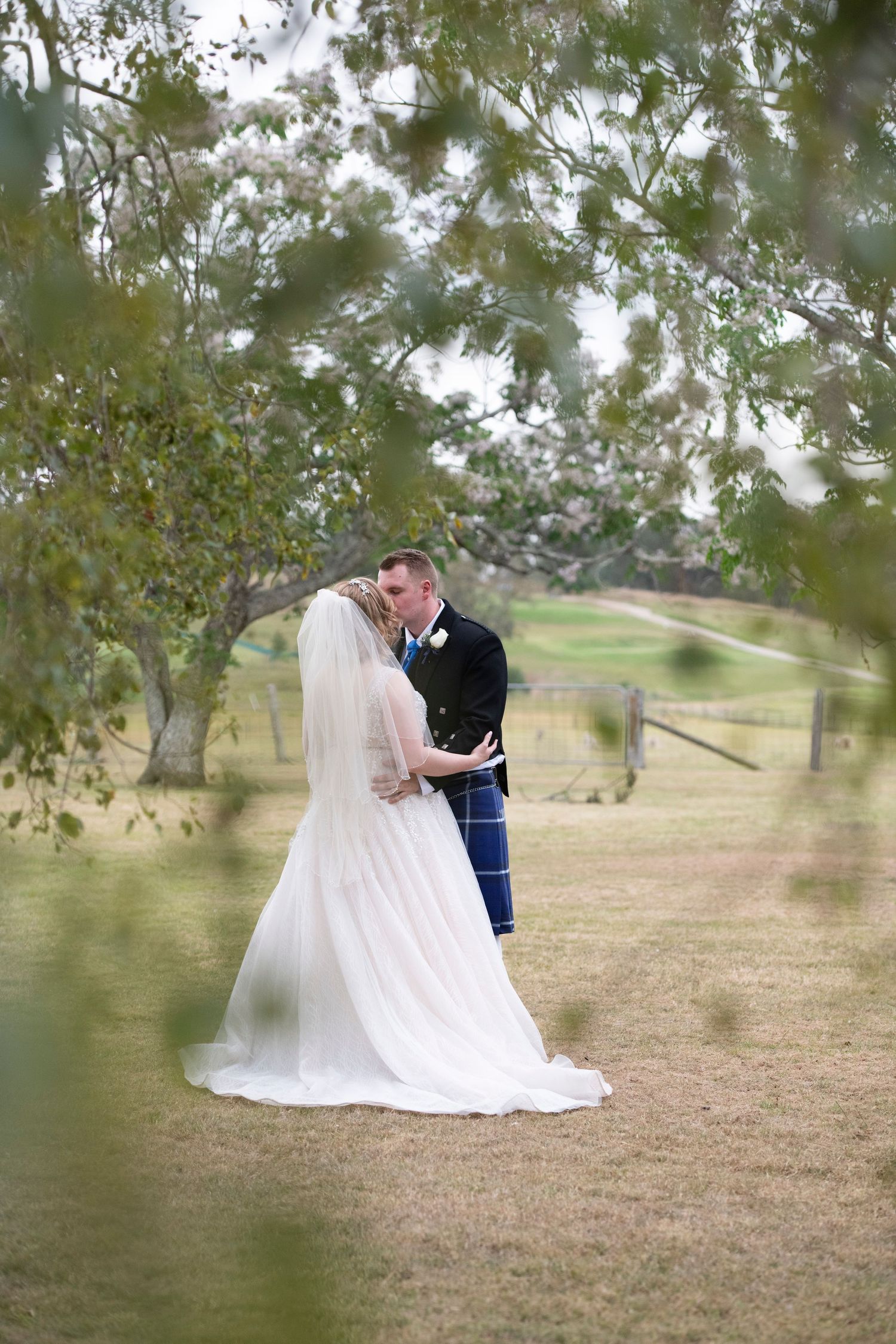 Bride and groom share a romantic kiss under lush green trees on a grassy lawn at an outdoor wedding venue.