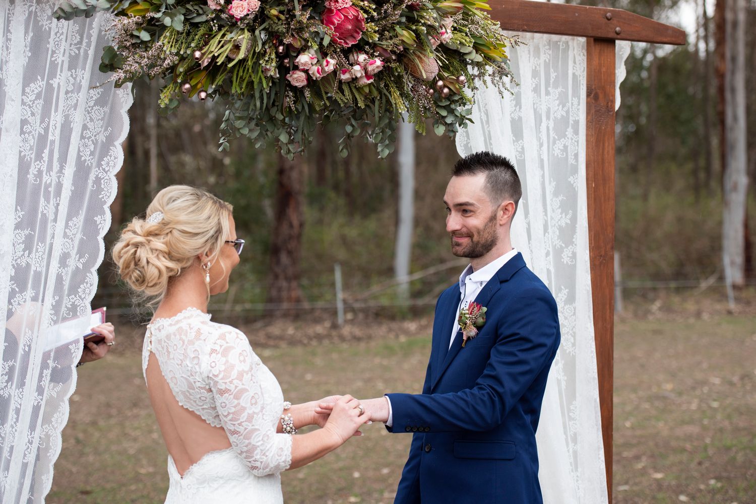 A wedding ceremony under a rustic wooden arch decorated with pink and native flowers and flowing white fabric.