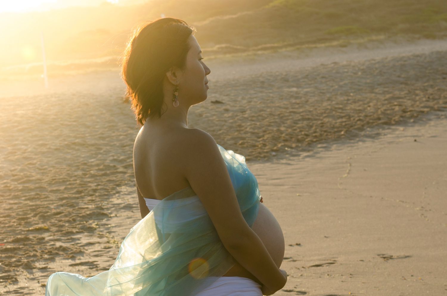 Silhouettes of mother to be walking along beach at sunset with waves in background.