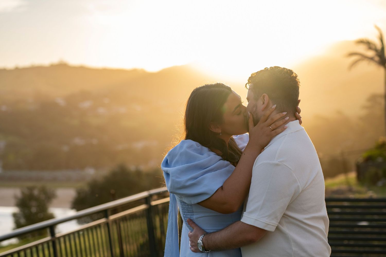 A couple embraces during golden hour sunset with palm trees silhouetted in the background.