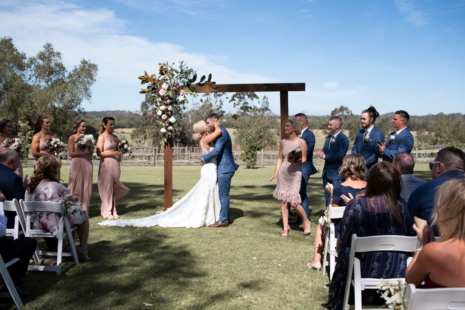Outdoor wedding ceremony setup with white chairs and guests under a bright blue sky.