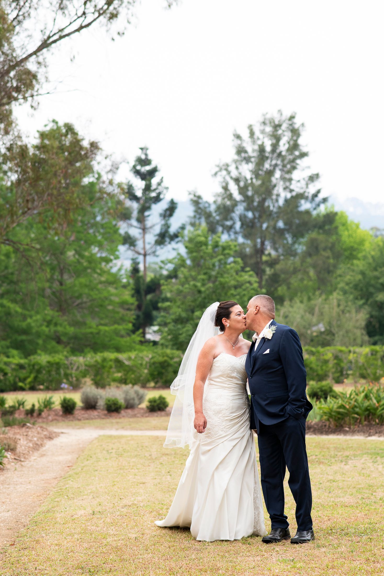 A romantic wedding couple shares tender kisses in a series of photos taken on a grassy field with trees in the background.