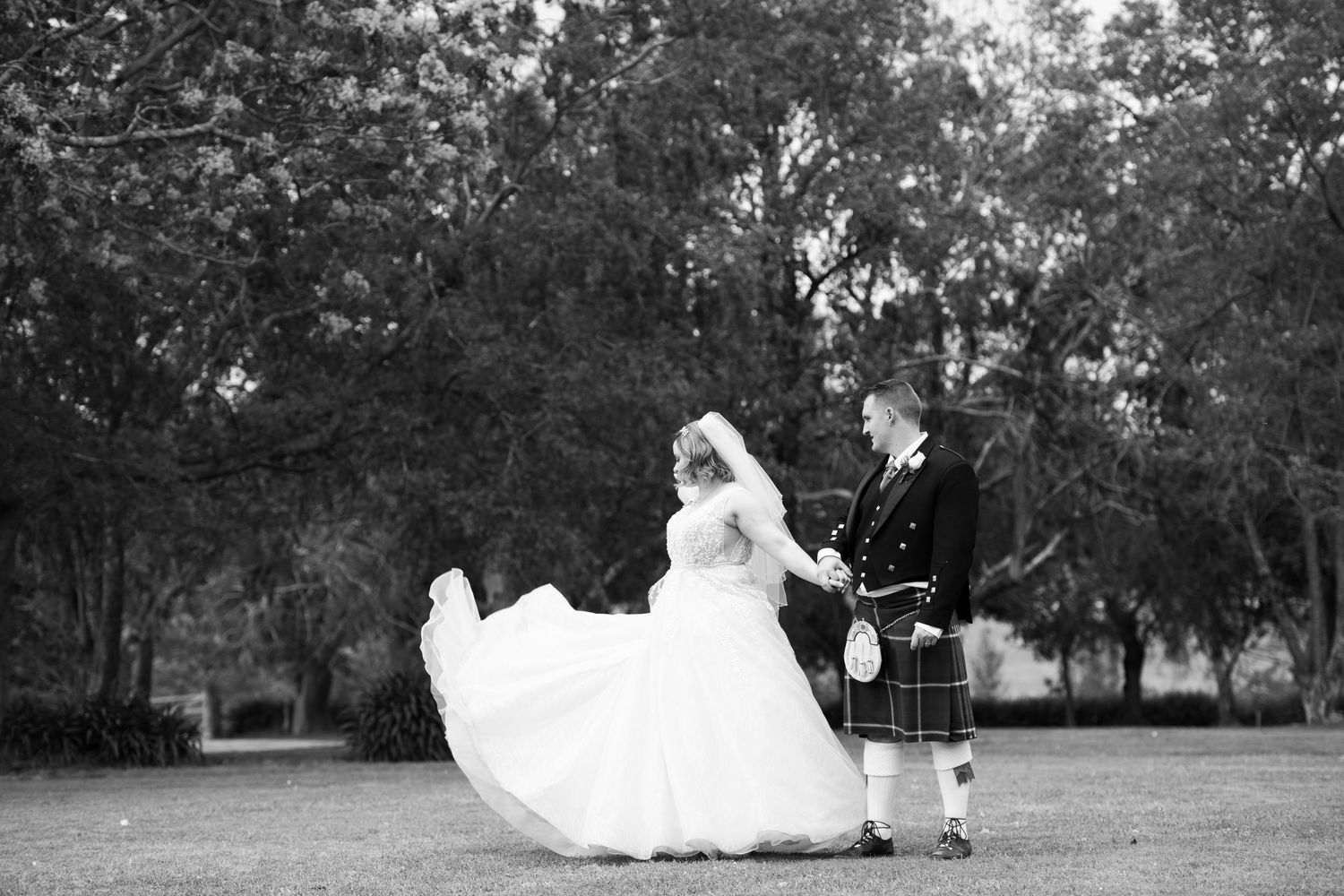 Black and white photo of a bride in flowing gown and groom in kilt dancing outdoors among trees.