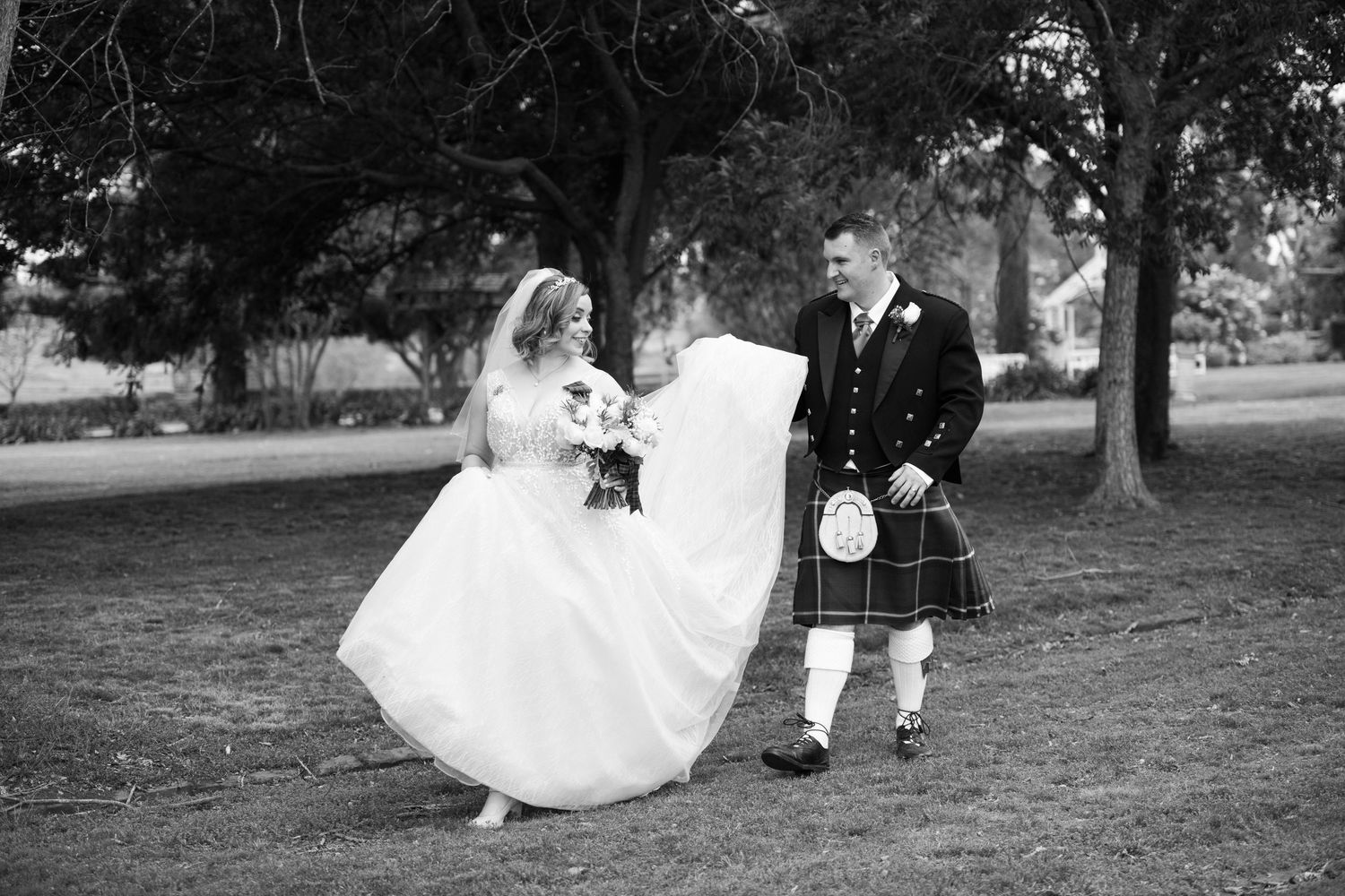 Black and white photo of a bride in a flowing gown and groom in a Scottish kilt walking together outdoors.