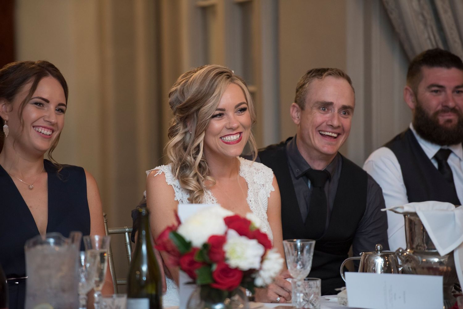 Wedding bride and groom with guests laugh and smile while seated at a long reception table decorated with red roses and wine glasses.