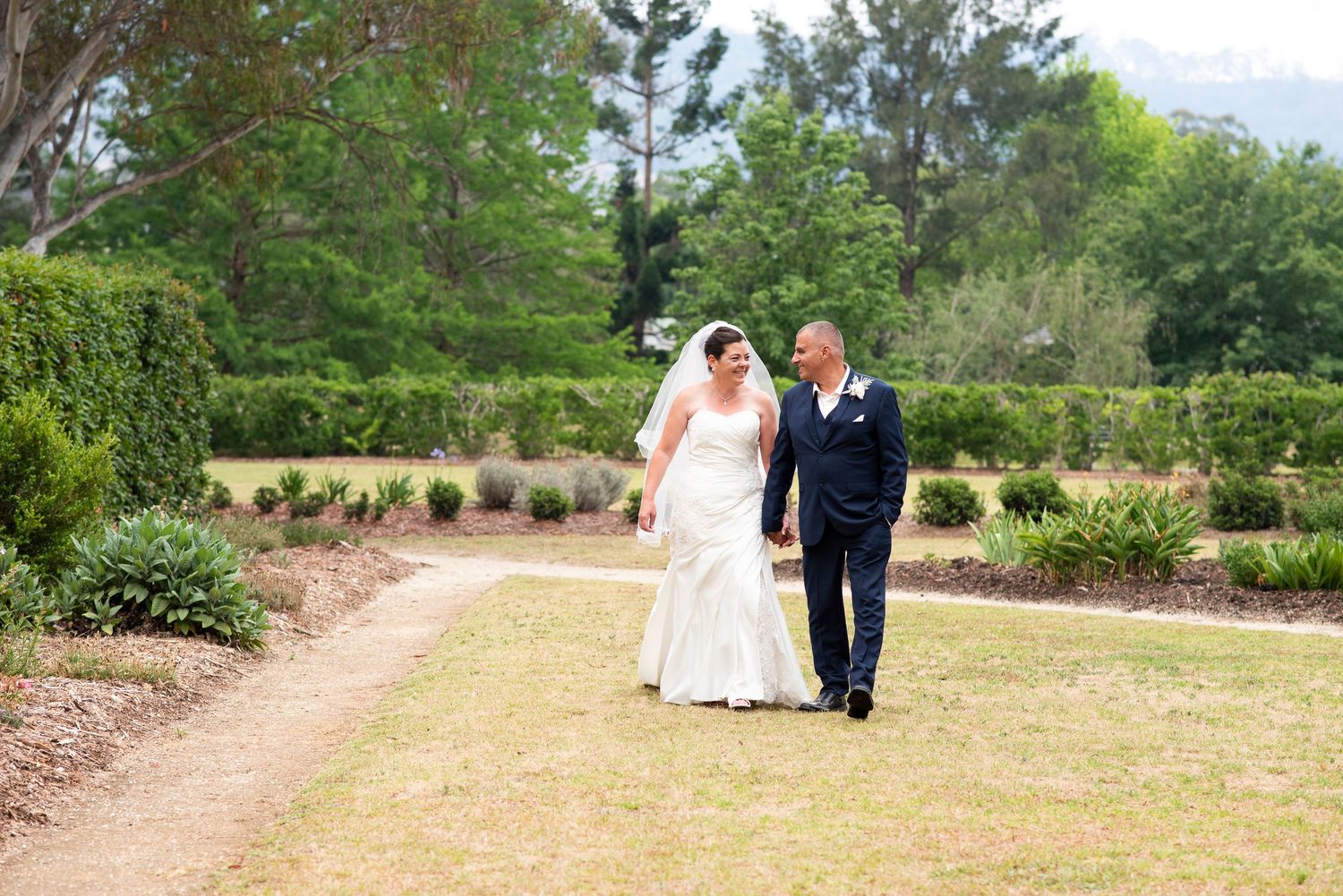 A couple in wedding attire walks along a scenic outdoor path surrounded by lush greenery and trees.