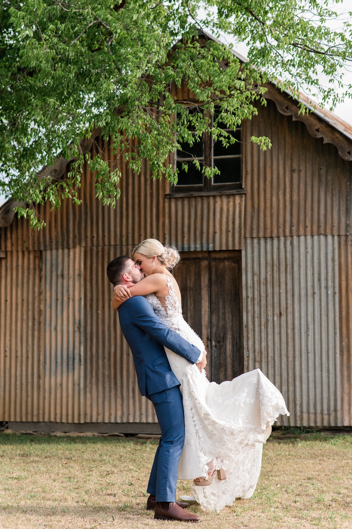 Bride and groom with romantic lift pose in front of a rustic barn with overhanging green trees.