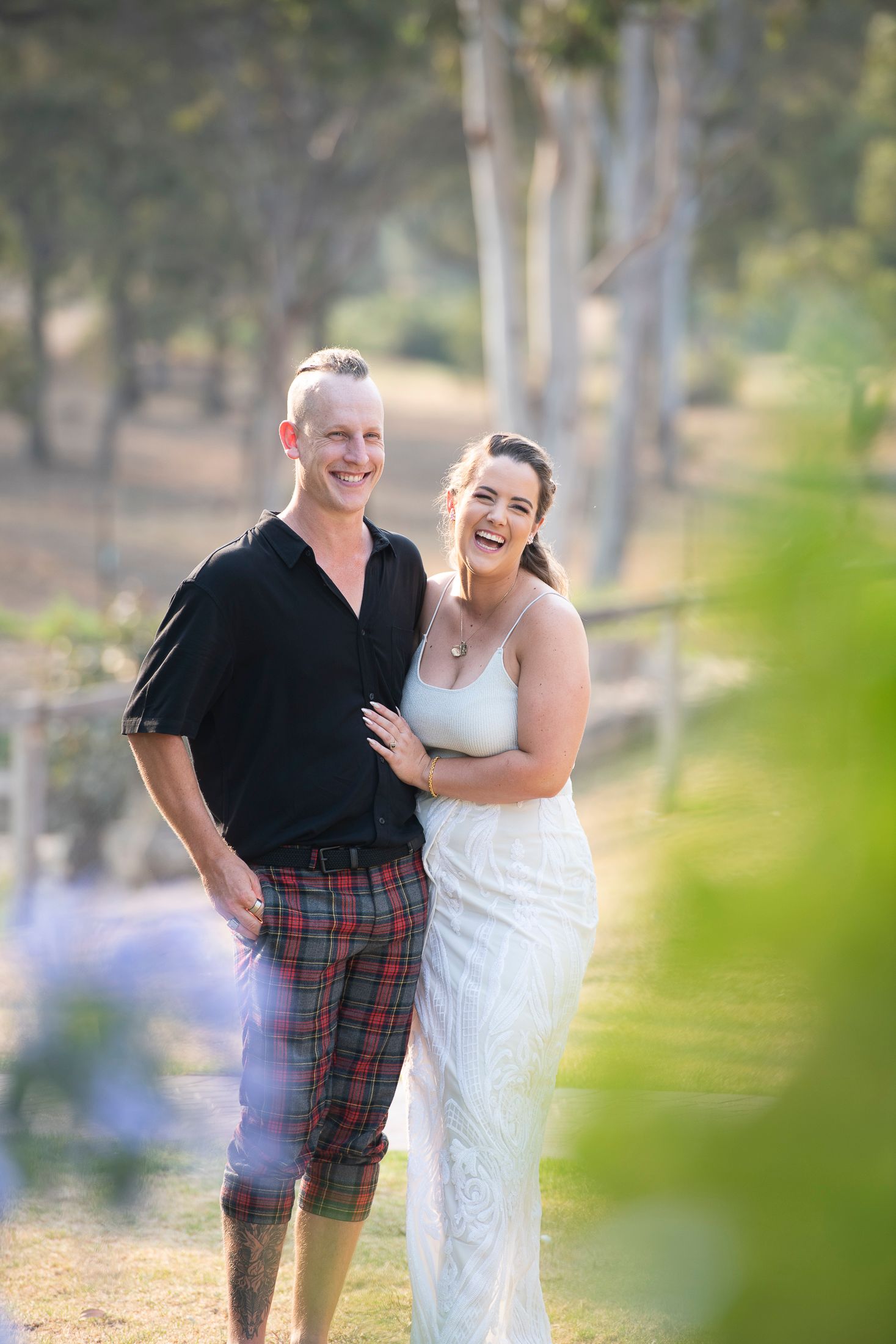 A couple shares joyful moments in a wooded outdoor setting wearing a white dress and black shirt with plaid kilt.