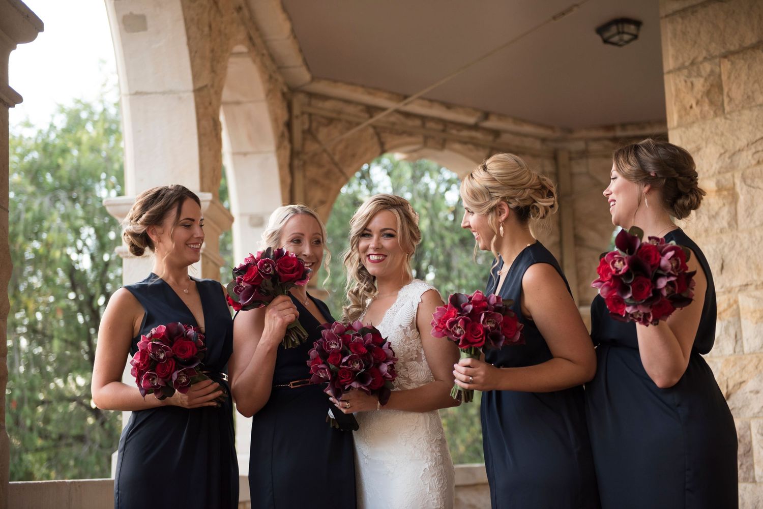 Bride with Bridesmaids in navy blue dresses hold deep red bouquets while standing under stone archways.