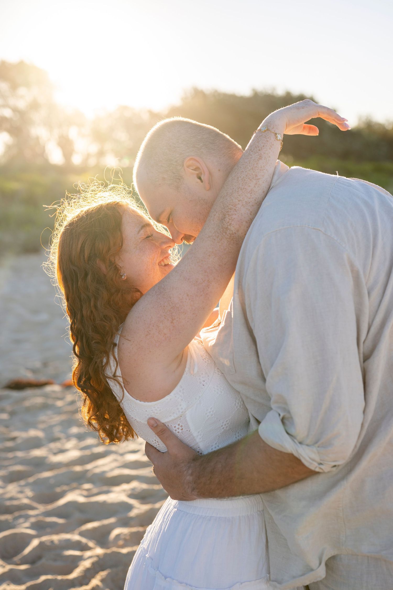 Couple embracing and almost kissing on a sandy beach at golden sunset, woman in white dress, man in linen shirt.