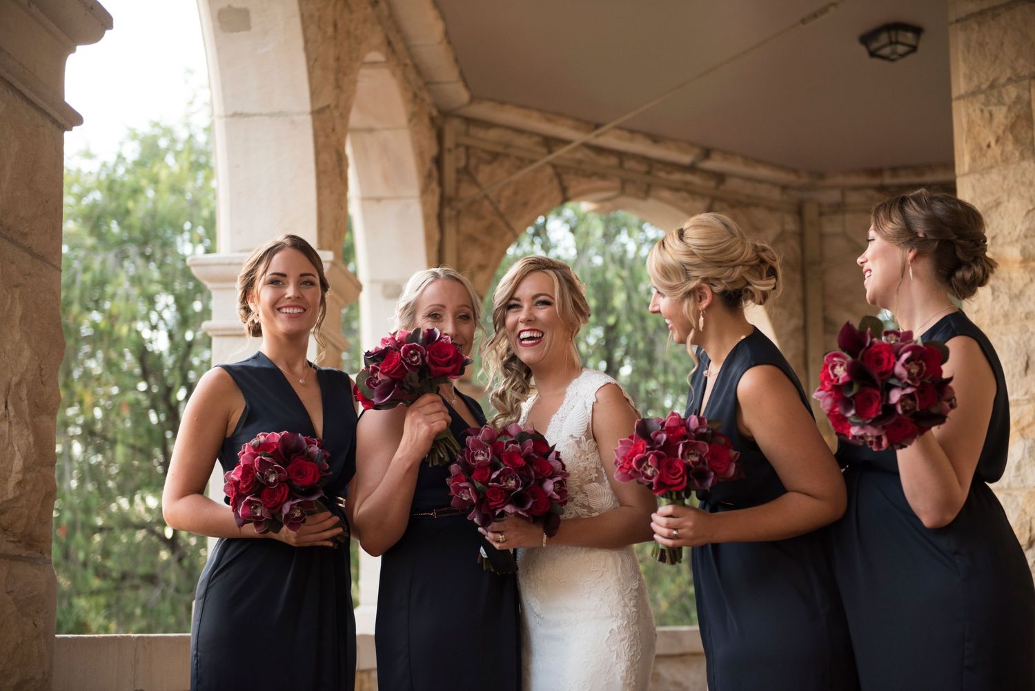 Brid with Bridesmaids in navy blue dresses holding red bouquets share joyful laughter during wedding photos under stone archway.